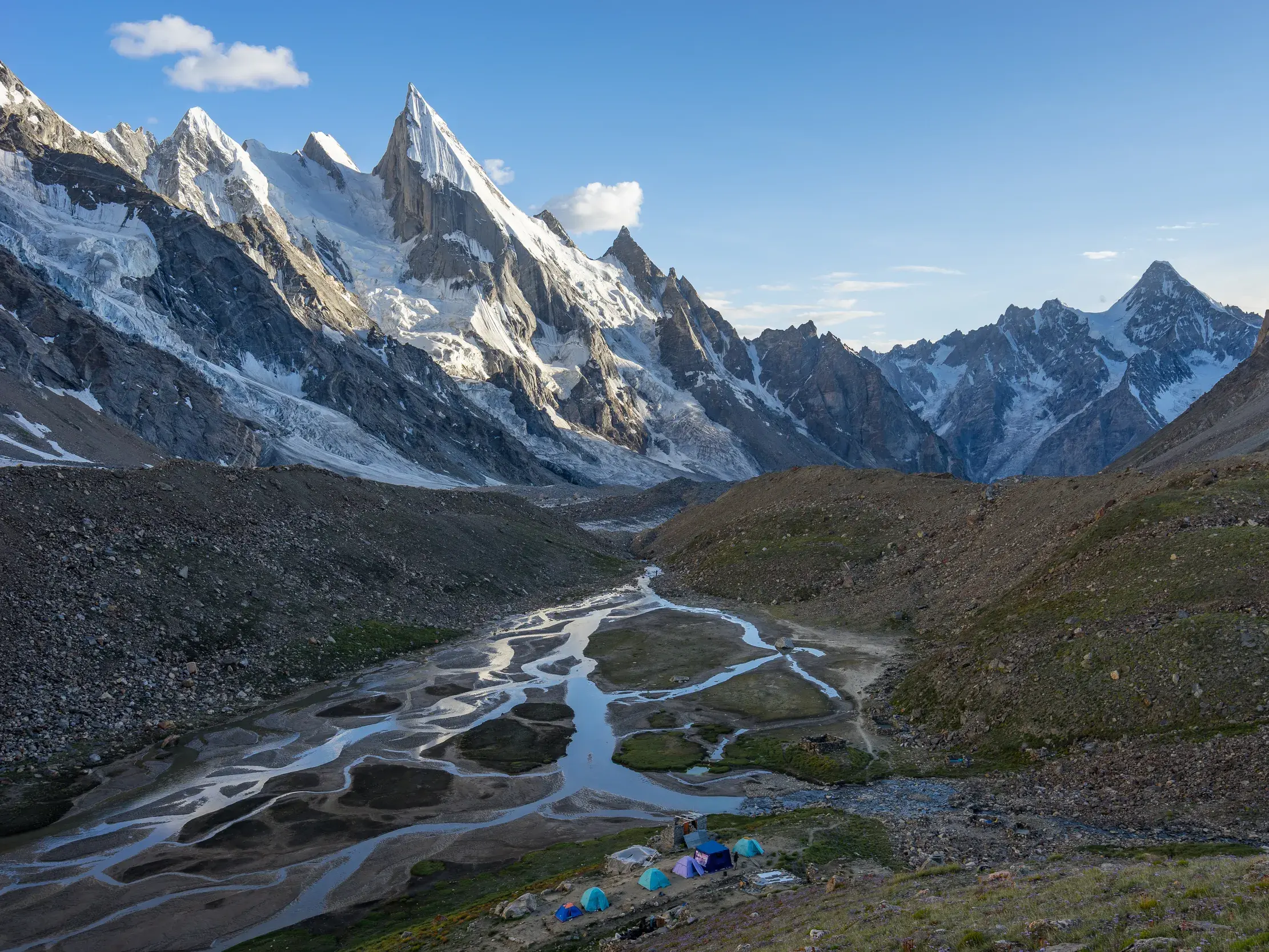 Zelte eines Trekkingcamps an einem Gletscherfluss im Hushe-Tal vor der imposanten, schneebedeckten Felsspitze des Laila Peak.