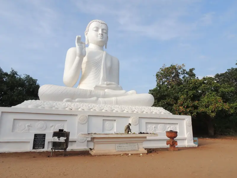 Große, sitzende Buddha-Statue in strahlendem Weiß auf dem heiligen Berg Mihintale, Sri Lanka, mit einem Affen davor.