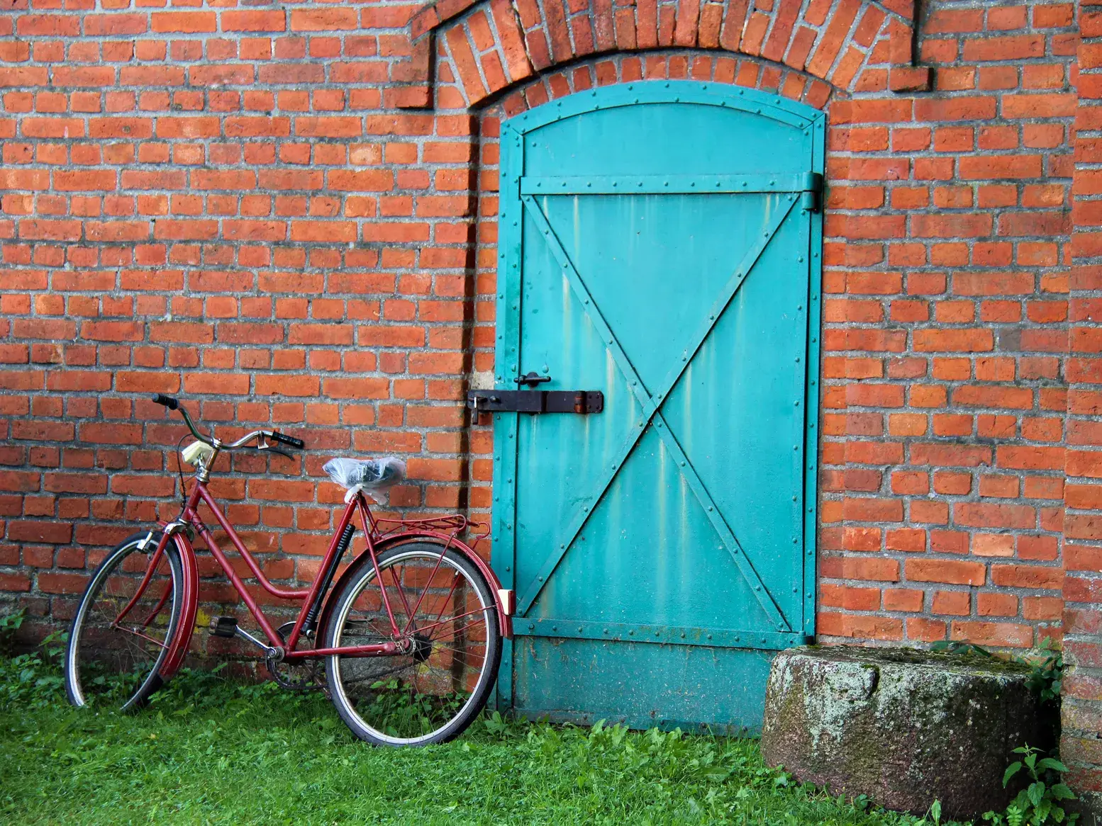 Rotes Fahrrad lehnt an einer rustikalen Backsteinwand neben einer türkisen Tür in einem ländlichen Dorf in Polen.