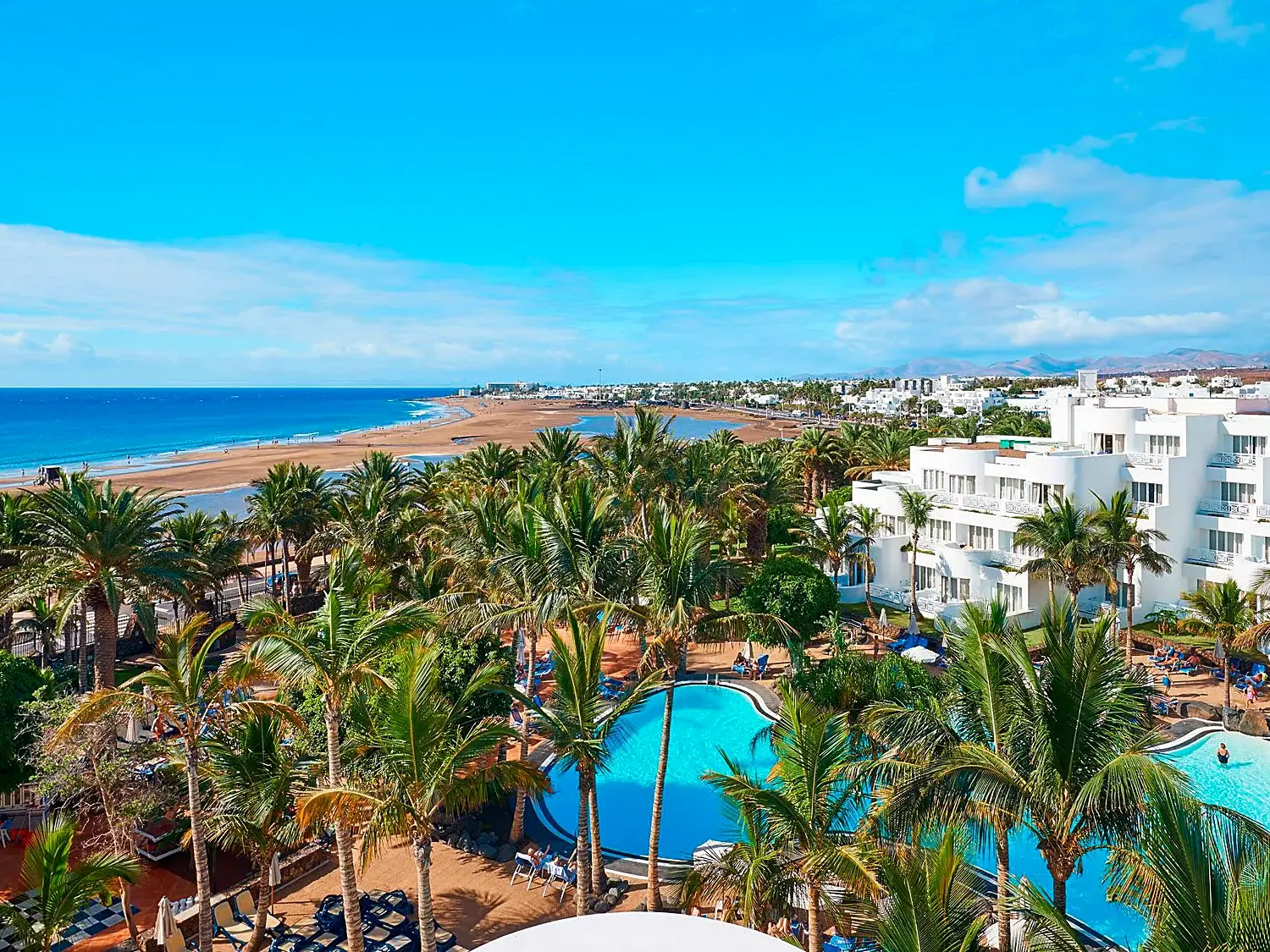 Blick von einem Hotel auf den Pool, Palmen und den weiten Sandstrand von Puerto del Carmen auf Lanzarote unter blauem Himmel.