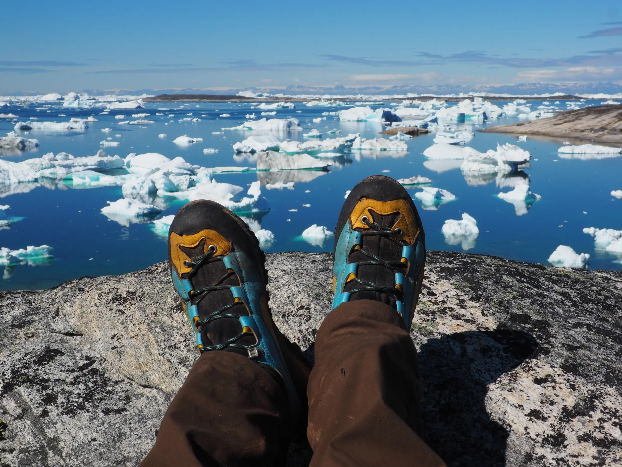 Ein Wanderer macht Rast auf einem Felsen und blickt aus der Ich-Perspektive auf die unzähligen Eisberge im Ilulissat-Eisfjord.