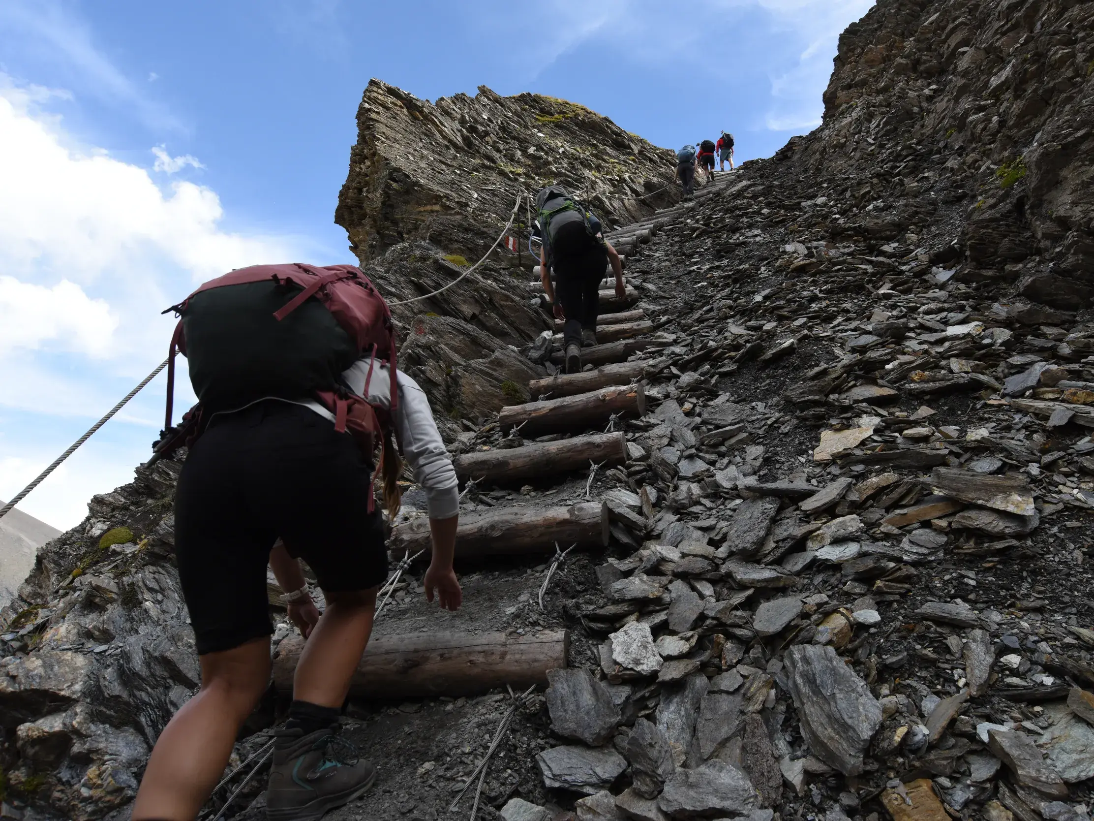 Eine Wandergruppe mit Rucksäcken steigt auf einer anspruchsvollen Etappe einer Alpenüberquerung einen steilen Geröllhang hinauf.