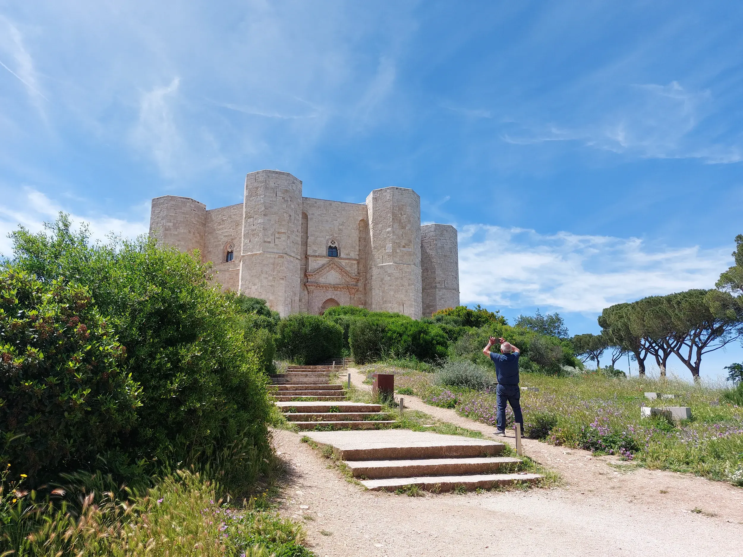 Ein Reisender fotografiert das achteckige Castel del Monte, das auf einem grünen Hügel in Apulien unter blauem Himmel thront.
