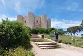 Ein Reisender fotografiert das achteckige Castel del Monte, das auf einem grünen Hügel in Apulien unter blauem Himmel thront.