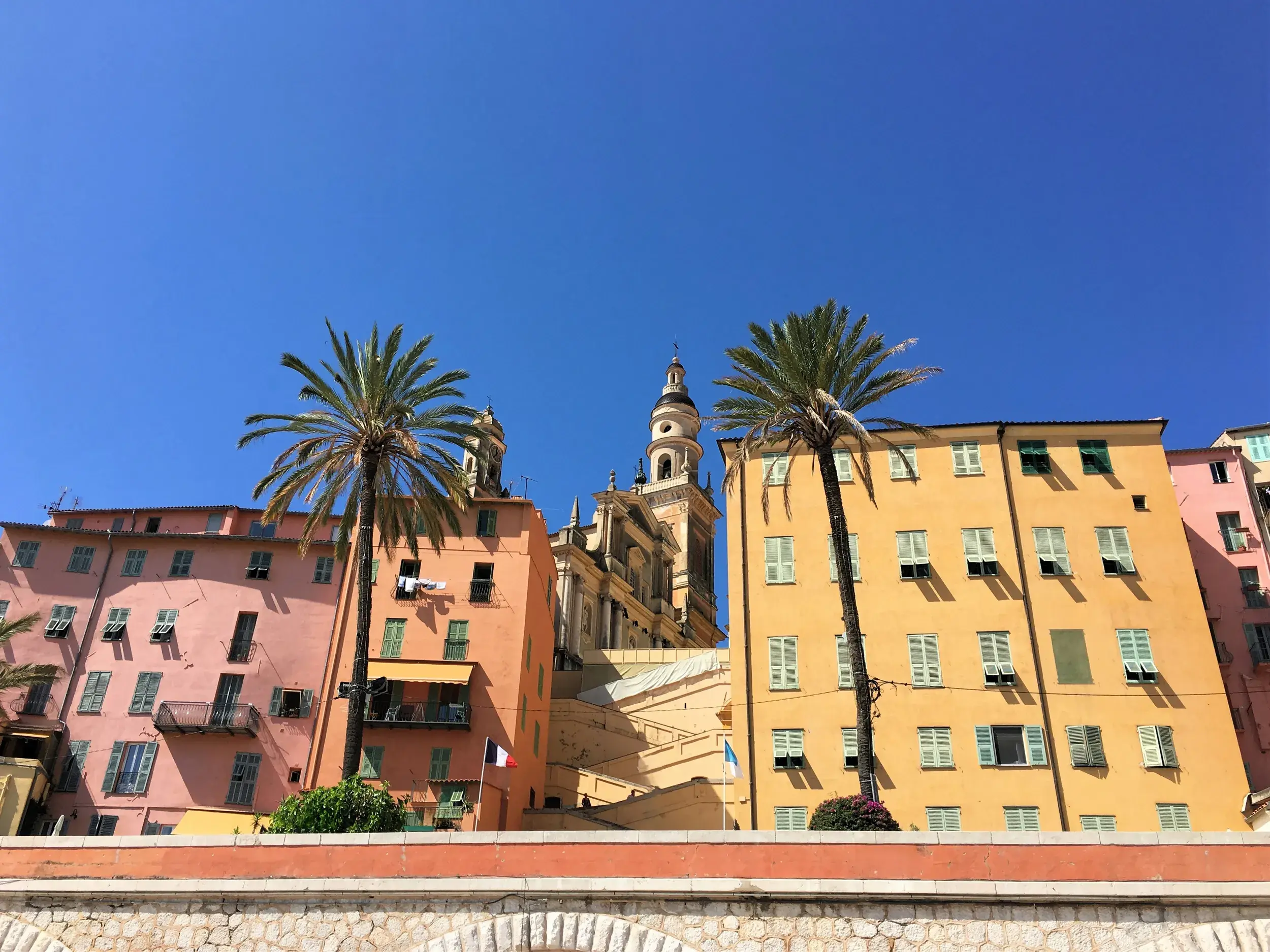 Sonniger Blick auf bunte Häuser und Palmen in der Altstadt von Menton mit der Basilika Saint-Michel-Archange im Hintergrund.