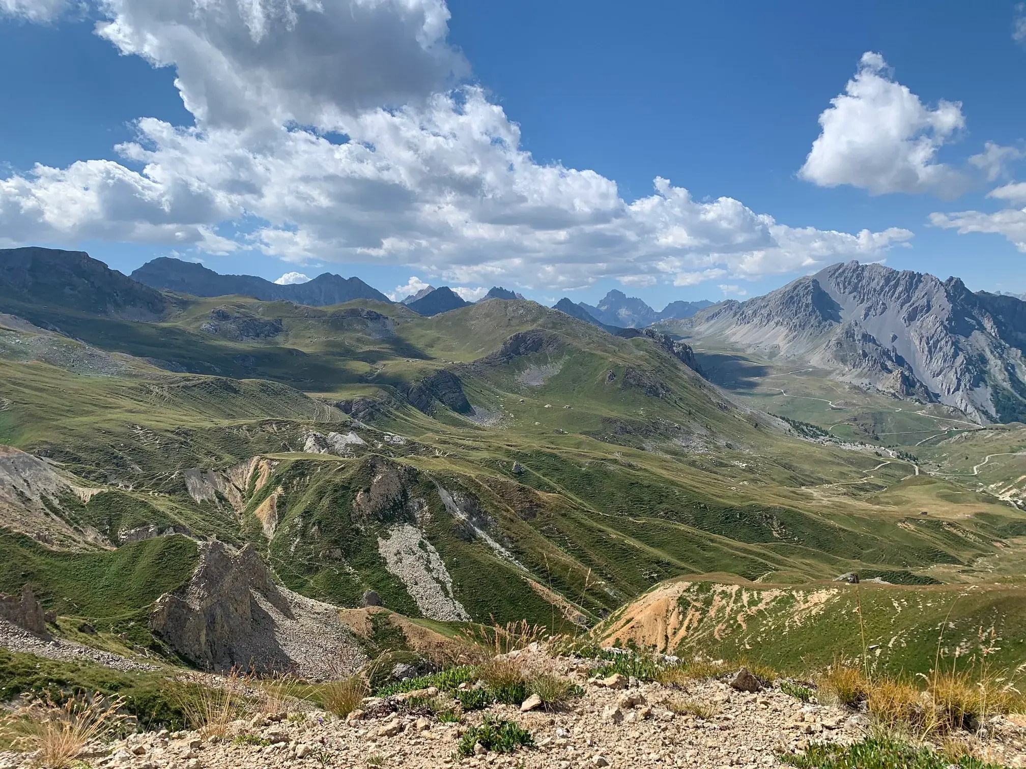 Weitblick beim Wandern über die grüne, hügelige Landschaft des Maira-Tals mit den felsigen Alpengipfeln im Hintergrund.