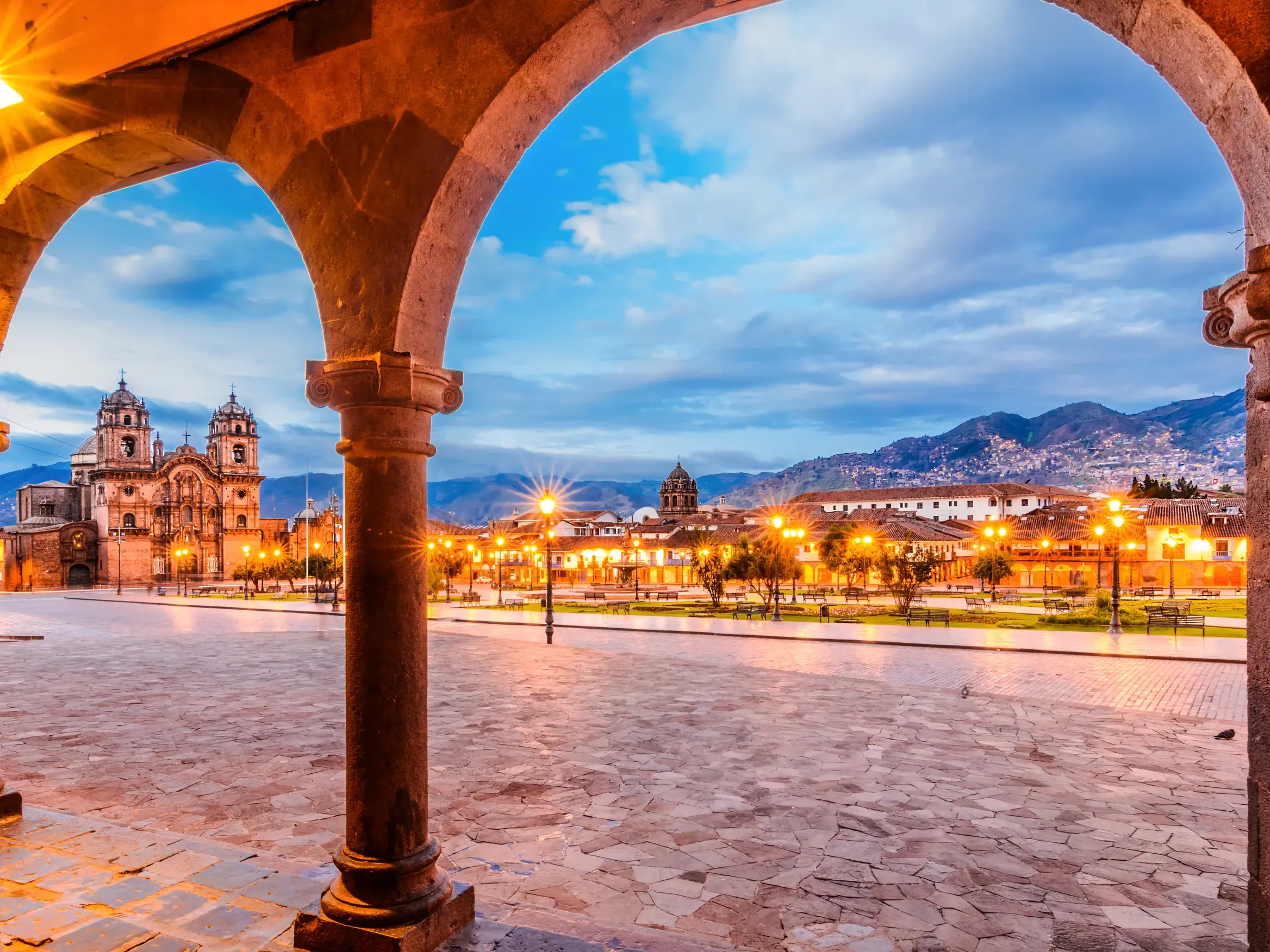Historische Arkaden rahmen den Blick auf den Plaza de Armas in Cusco mit der Jesuitenkirche, die bei Dämmerung vor den Anden leuchtet.