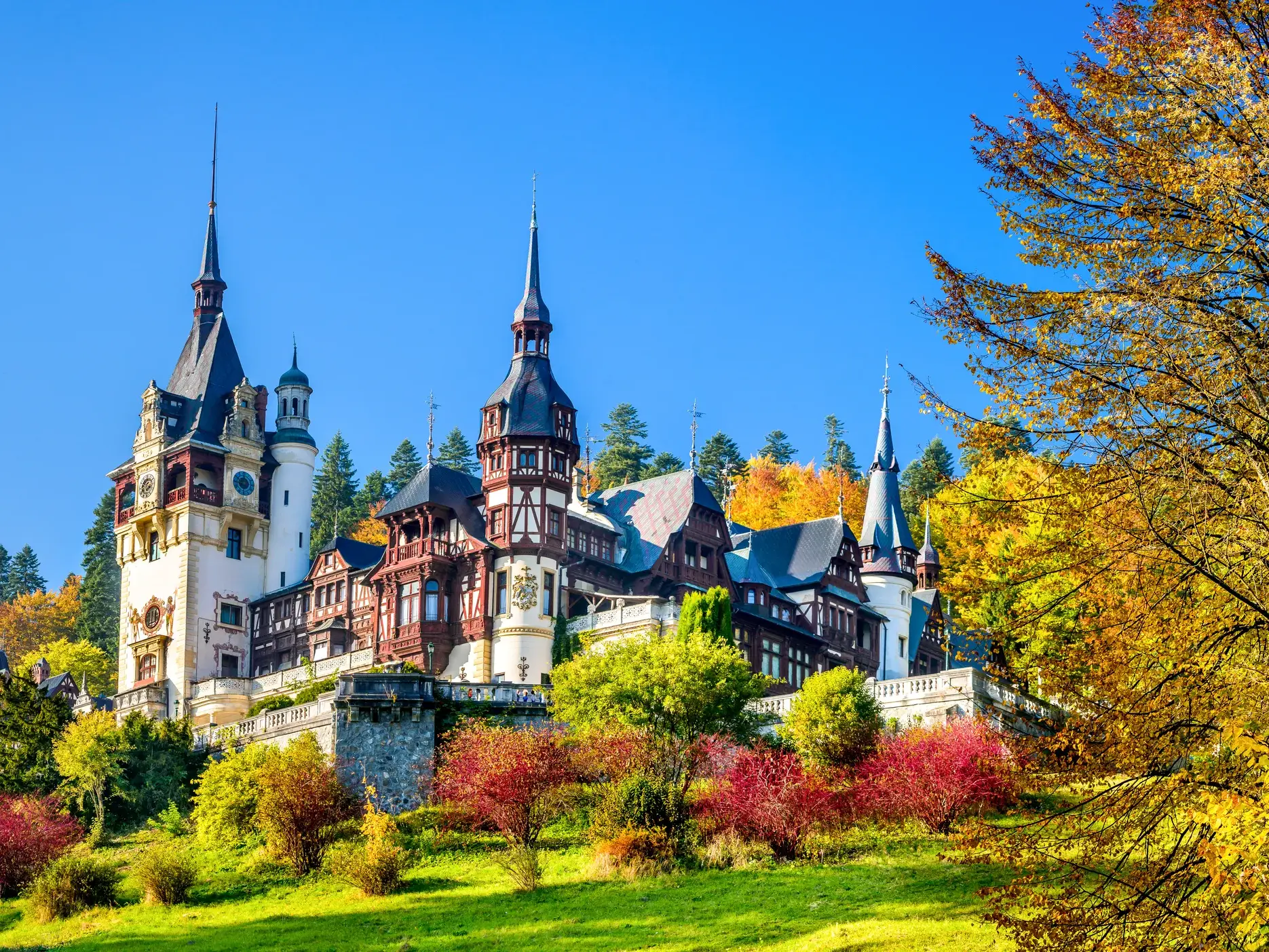 Das märchenhafte Schloss Peles in Rumänien, eingebettet in herbstlich gefärbte Wälder der Karpaten bei strahlendem Sonnenschein.