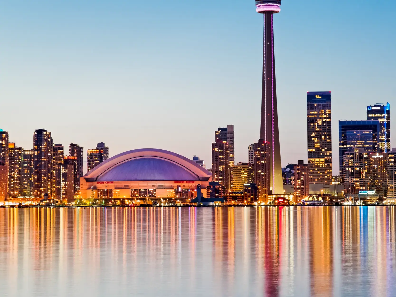 Die beleuchtete Skyline von Toronto mit dem CN Tower und Rogers Centre spiegelt sich bei Dämmerung im Wasser des Ontariosees.