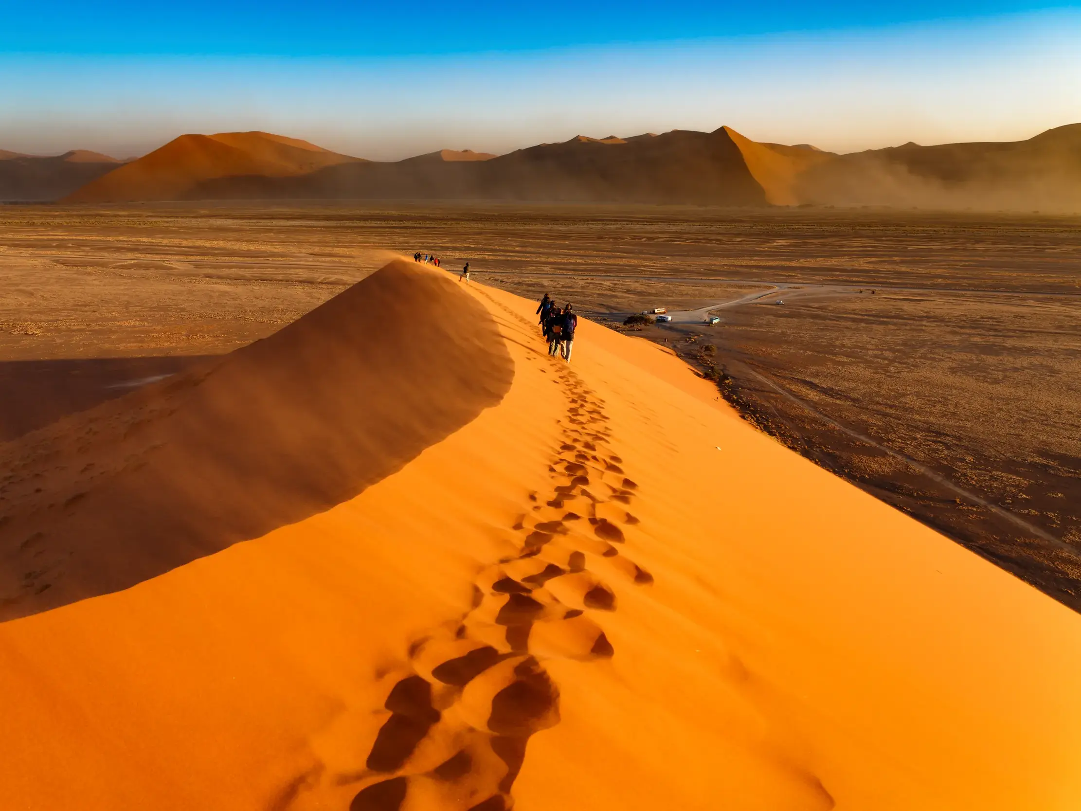 Eine kleine Reisegruppe besteigt den schmalen Grat einer riesigen, orangefarbenen Sanddüne im Sossusvlei in Namibia.