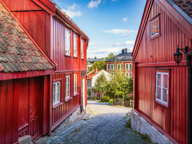 Malerische Kopfsteinpflastergasse in Damstredet, Oslo, gesäumt von historischen, leuchtend roten Holzhäusern an einem sonnigen Tag.