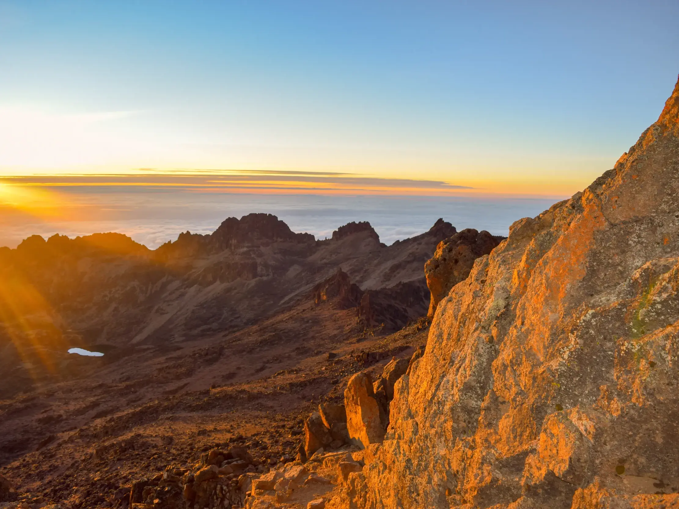 Goldener Sonnenaufgang vom Gipfel des Point Lenana am Mount Kenya mit Blick auf schroffe Felsen und den Lake Michaelson.