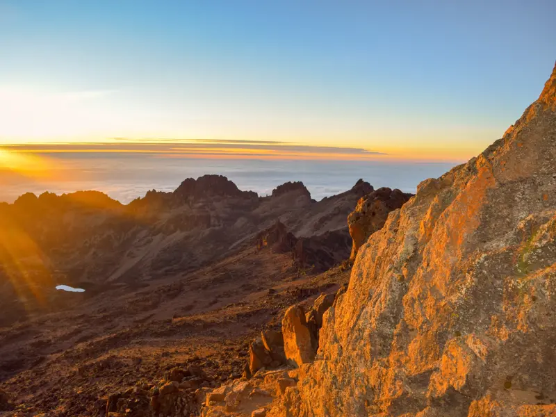Goldener Sonnenaufgang vom Gipfel des Point Lenana am Mount Kenya mit Blick auf schroffe Felsen und den Lake Michaelson.