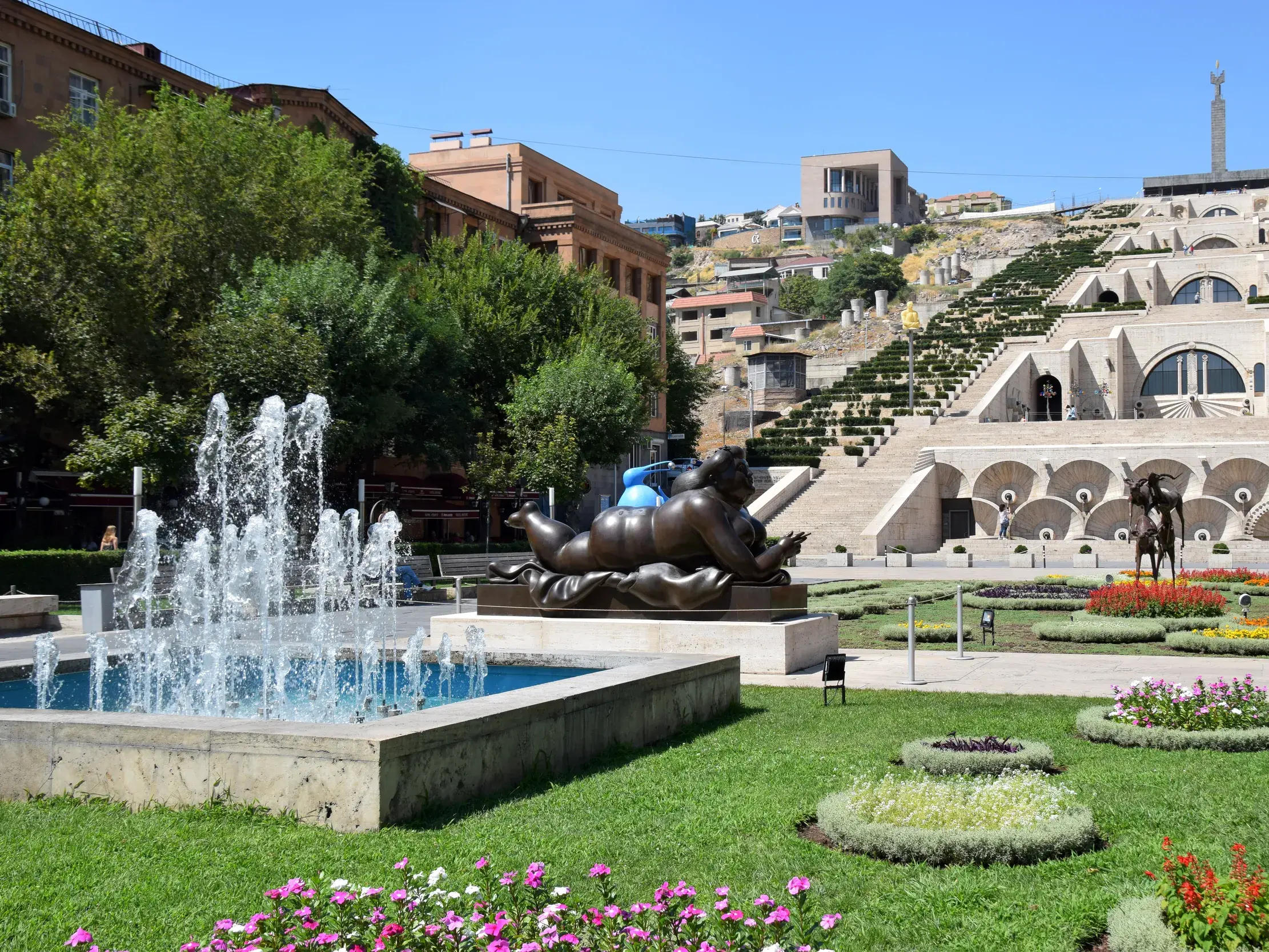 Sonniger Blick auf die Kaskaden in Eriwan mit einem Park, Springbrunnen und einer modernen Skulptur im Vordergrund.