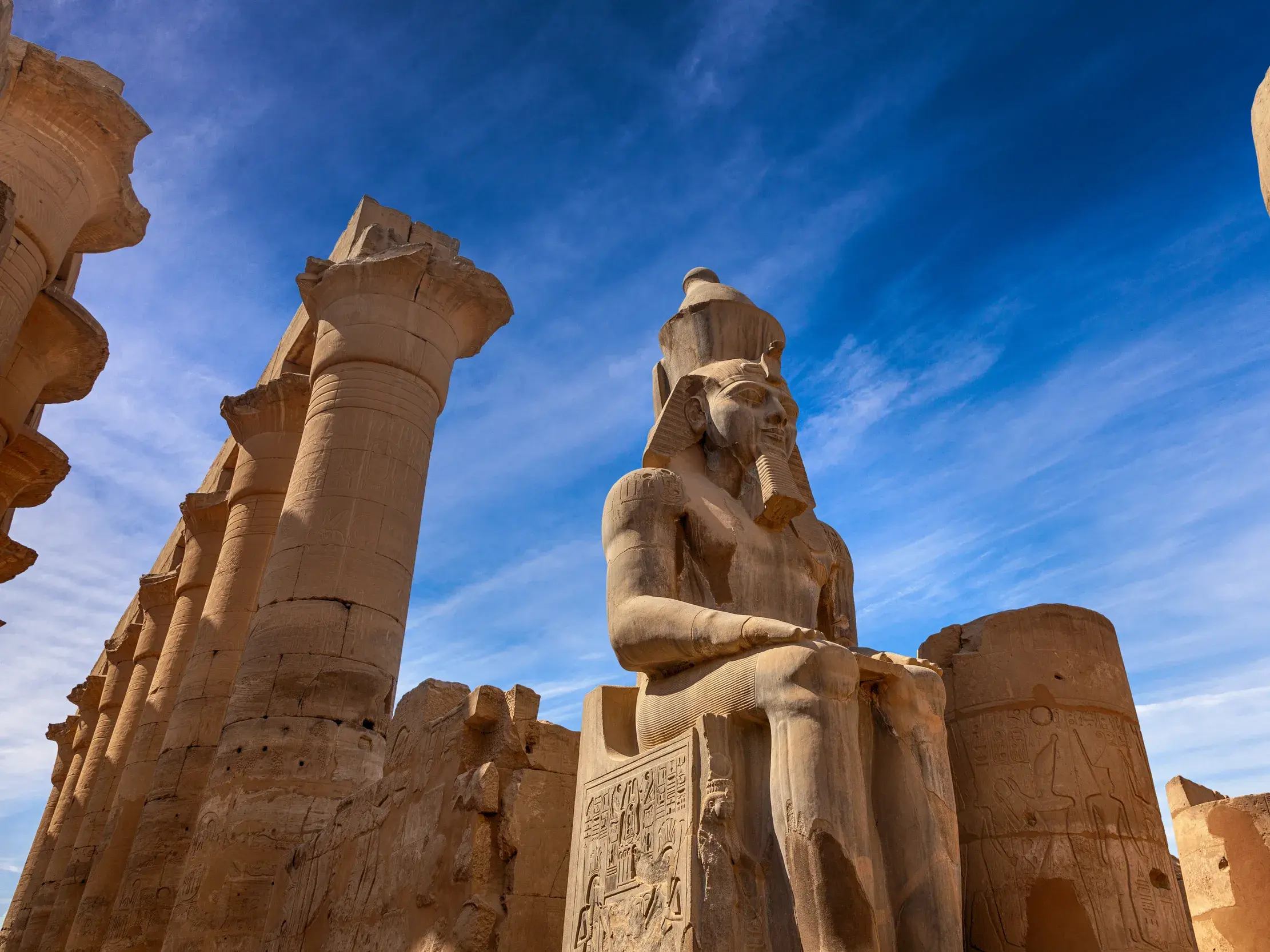 Majestätische Statue von Pharao Ramses II. auf einem Thron im Luxor-Tempel, umgeben von Säulen unter strahlend blauem Himmel.