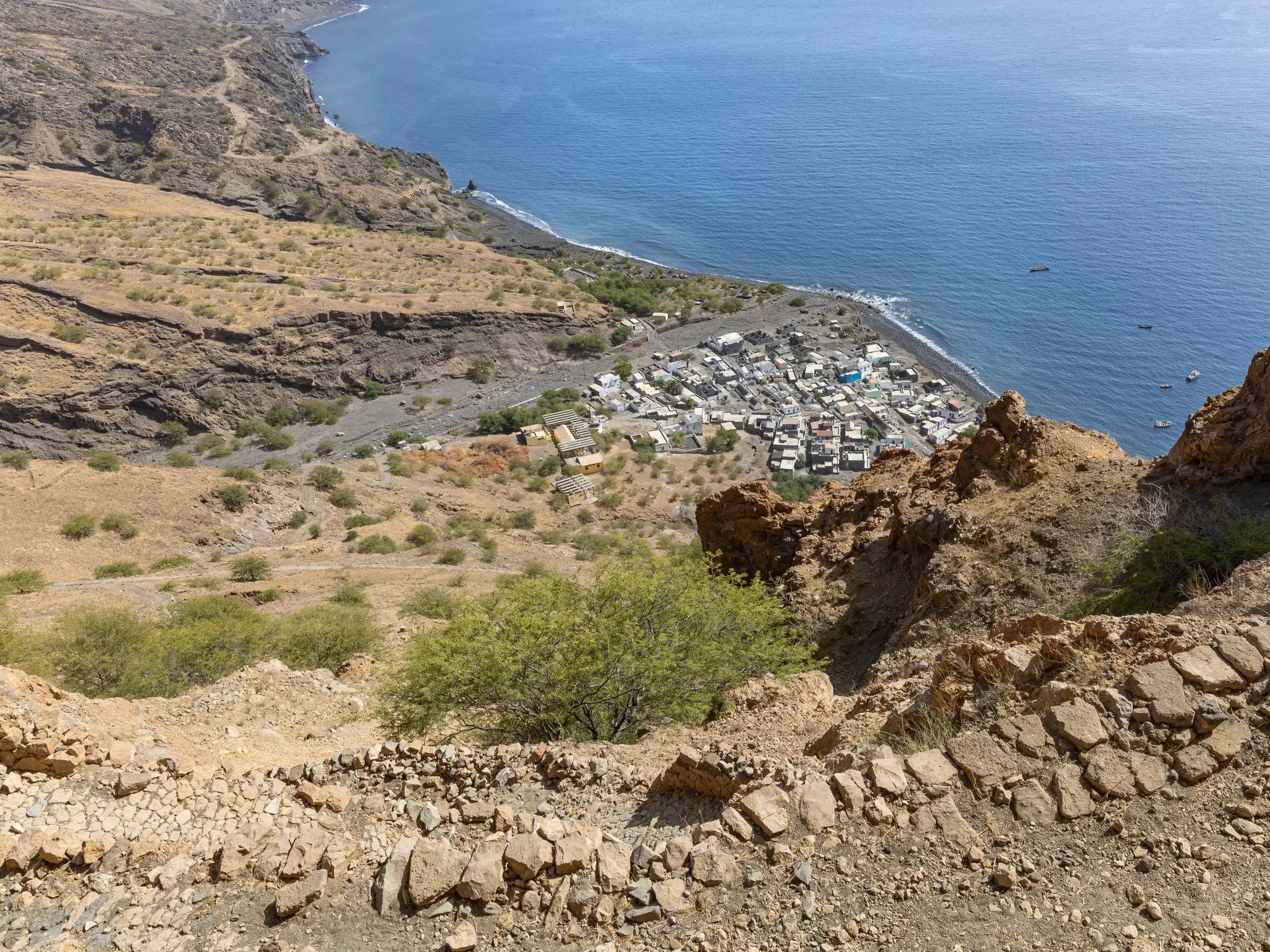 Ein steiniger Wanderpfad führt hinab zum Fischerdorf Monte Trigo auf Santo Antão, umgeben von kargen Hängen und dem Atlantik.