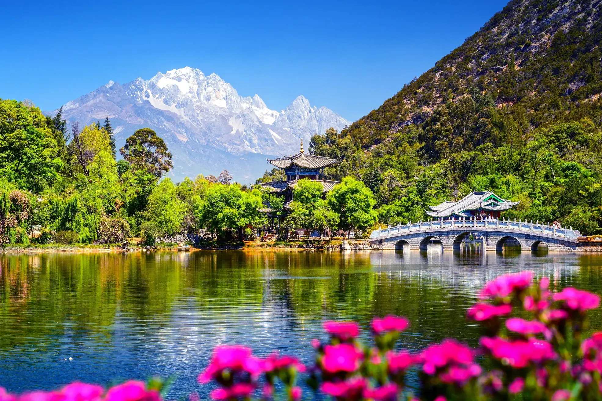 Idyllischer Schwarzer Drachen See in Lijiang mit einer Bogenbrücke und Pagode vor der Kulisse des Jade-Drachen-Schneebergs.
