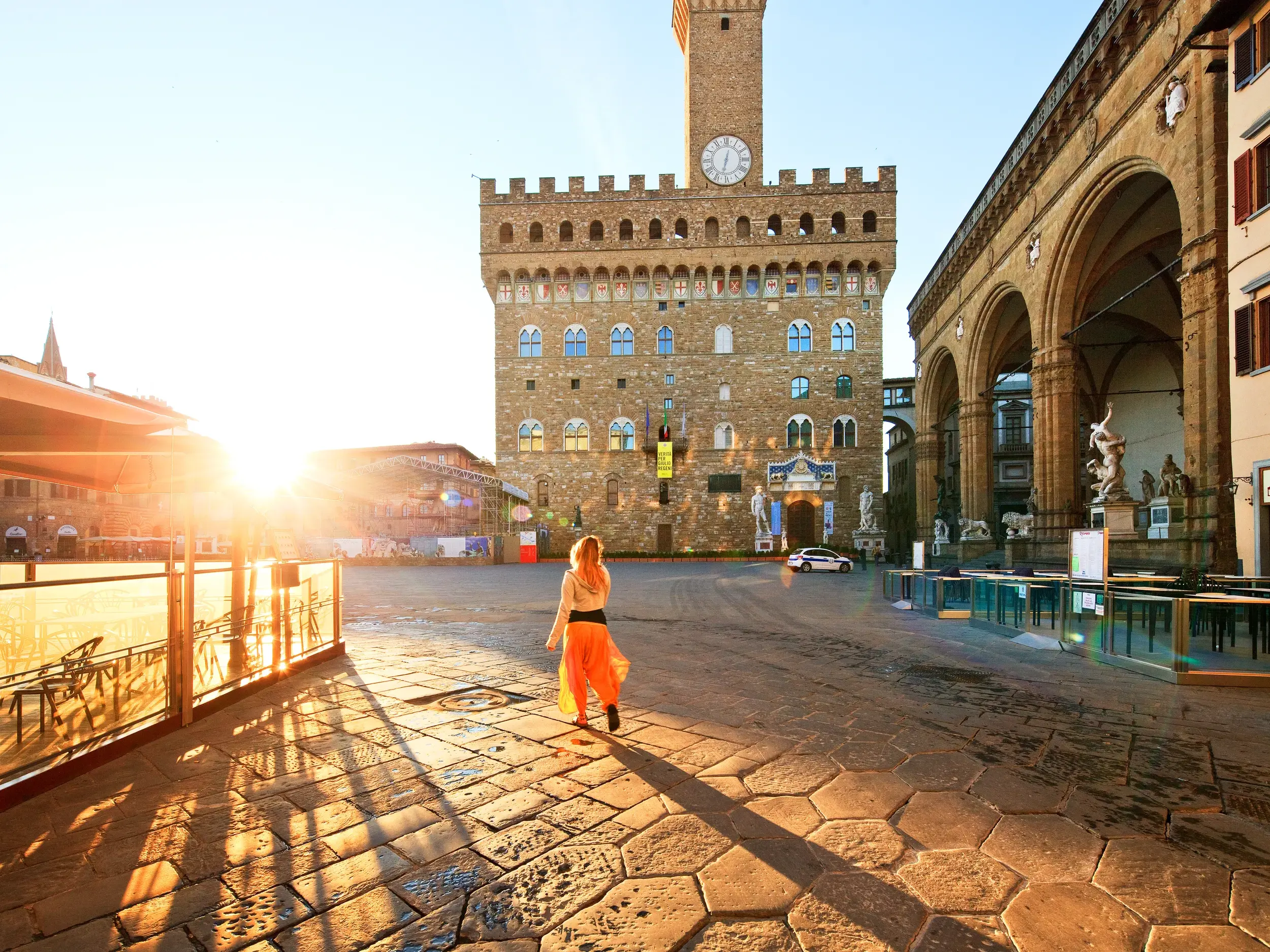 Eine Frau im orangefarbenen Rock erkundet im warmen Morgenlicht die Piazza della Signoria in Florenz vor dem Palazzo Vecchio.