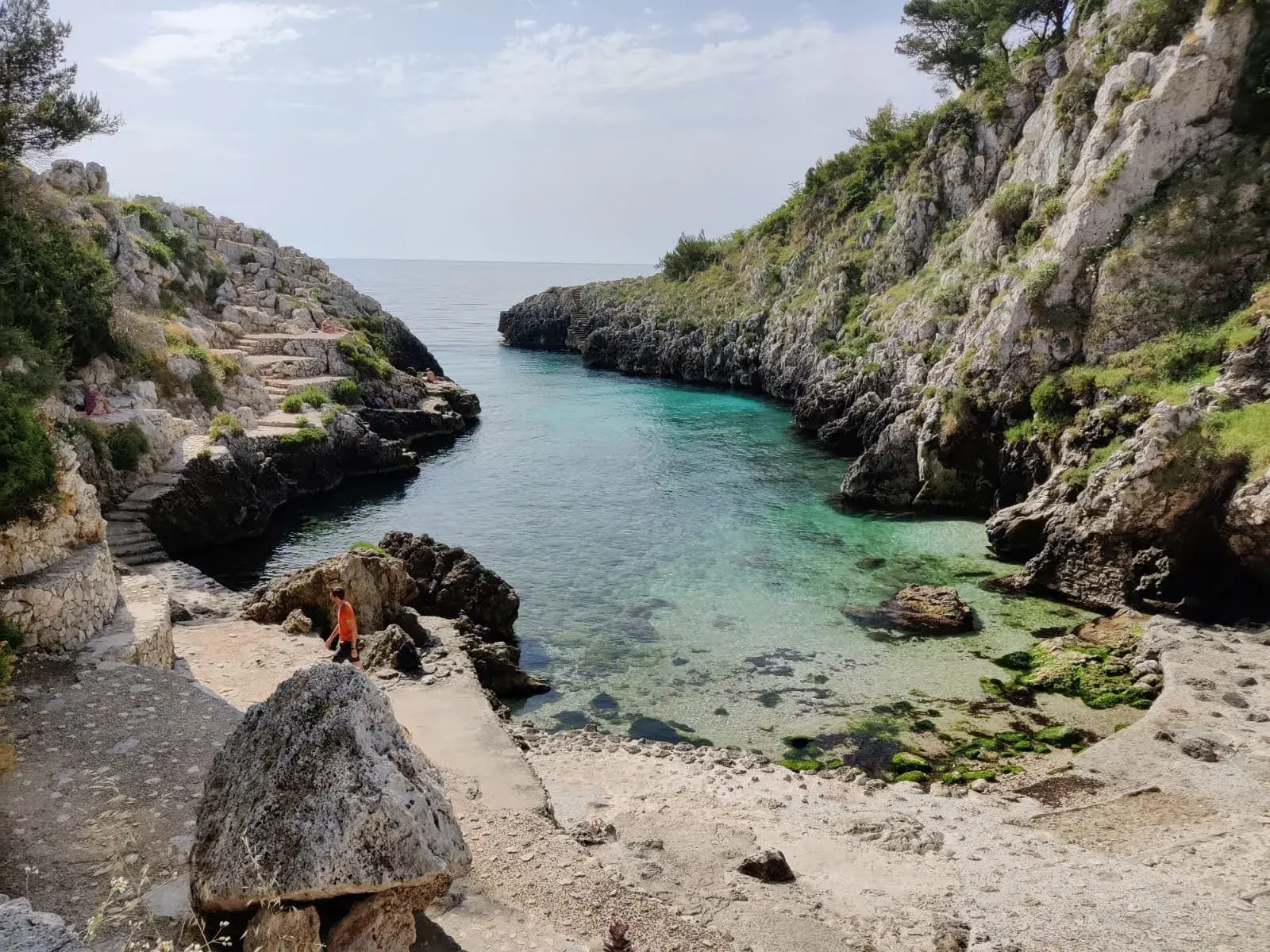 Felsige Bucht Cala dell'Acquaviva in Apulien mit klarem, türkisen Meer, Steintreppen und einem Wanderer am felsigen Ufer.