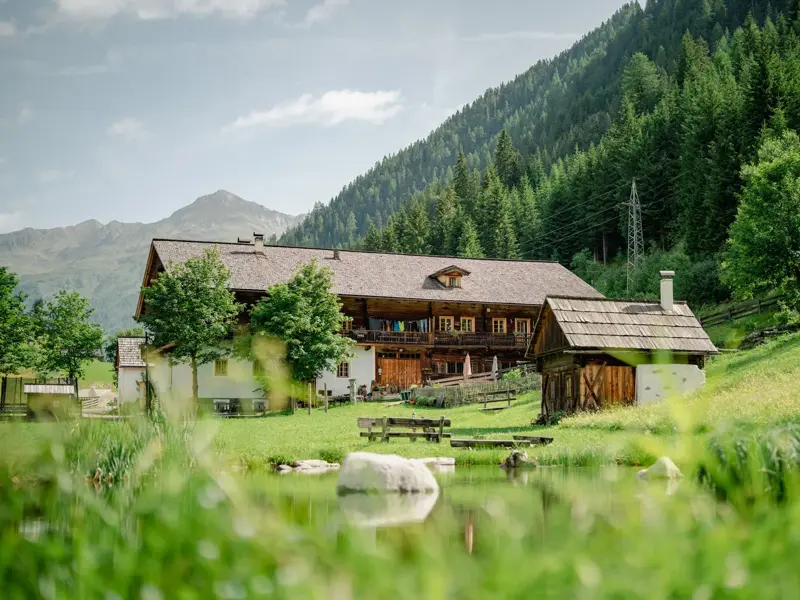 Der historische Wurzerhof im Villgratental, umgeben von Almwiesen und einem Teich, am Fuße eines bewaldeten Berges.