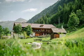 Der historische Wurzerhof im Villgratental, umgeben von Almwiesen und einem Teich, am Fuße eines bewaldeten Berges.