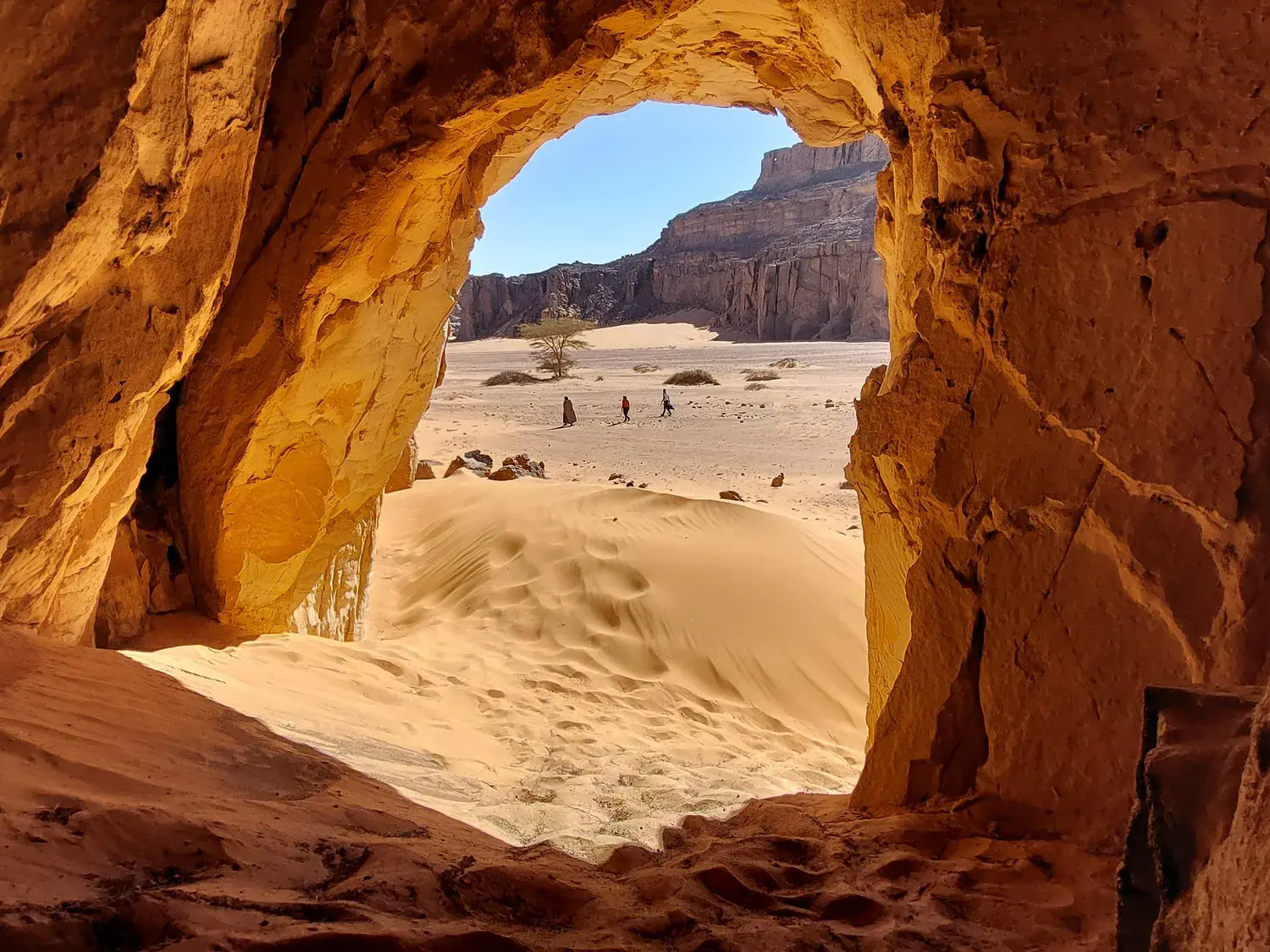 Aus einer Sandsteinhöhle blickt man auf Wanderer in der weiten Dünenlandschaft des Tassili n'Ajjer Nationalparks in Algerien.