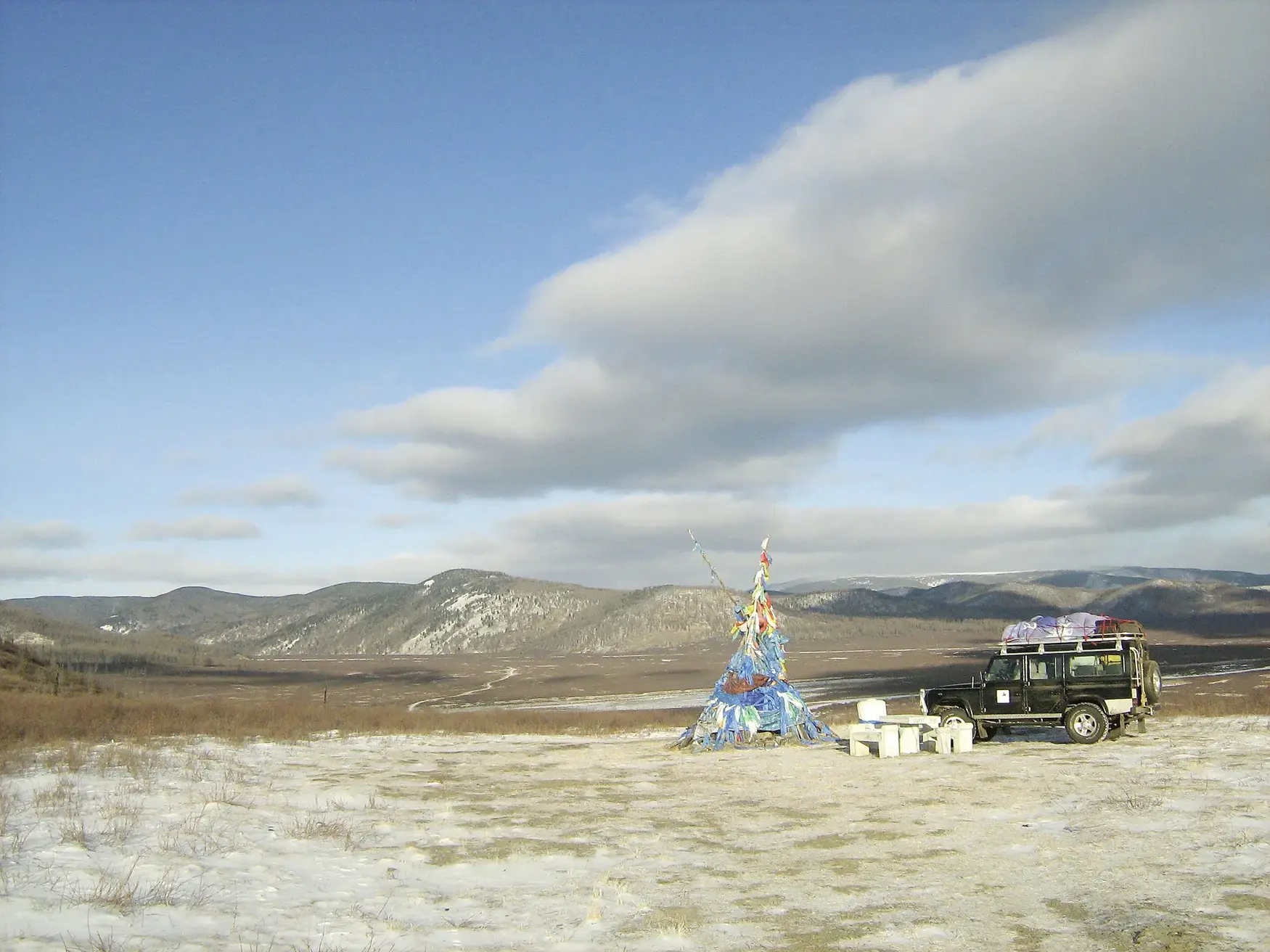 Begleitfahrzeug einer Radreise macht Rast an einem Ovoo mit Gebetsfahnen in der winterlichen Weite der mongolischen Steppe.