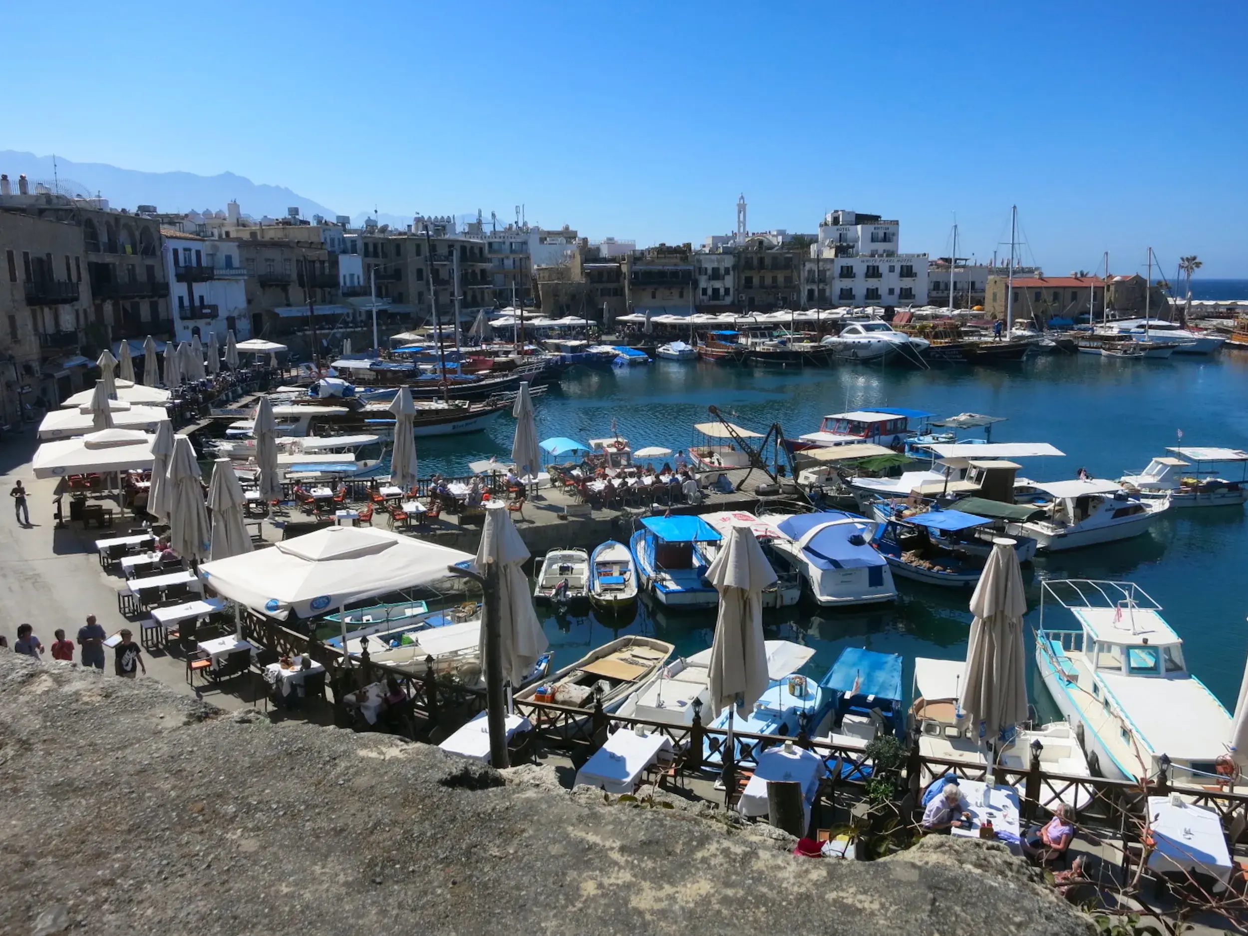 Sonniger Panoramablick auf den hufeisenförmigen Hafen von Kyrenia mit zahlreichen Booten und belebten Tavernen entlang der Uferpromenade.