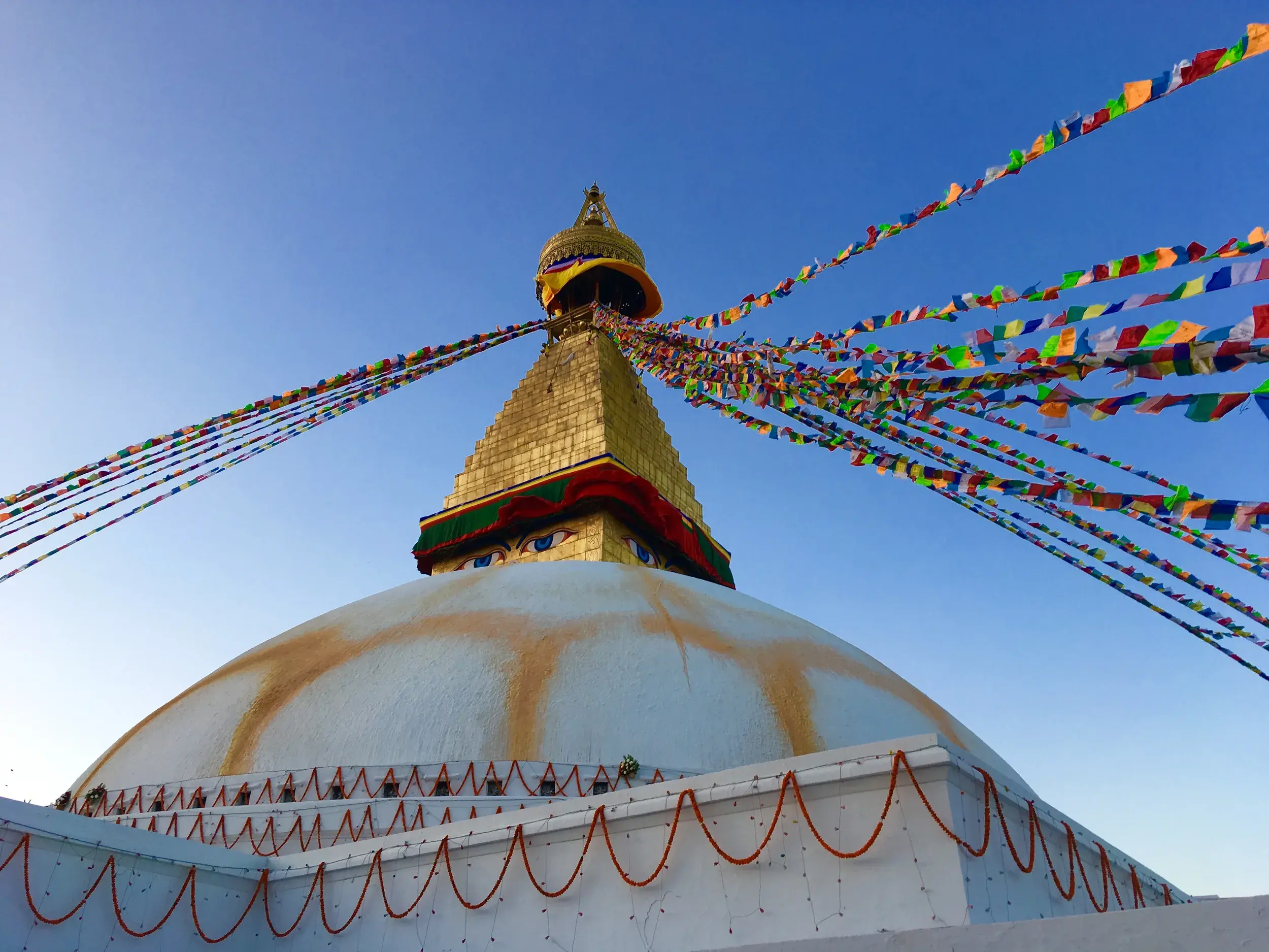 Die weiße Kuppel und der goldene Turm der Boudhanath Stupa in Kathmandu, geschmückt mit bunten Gebetsfahnen vor blauem Himmel.