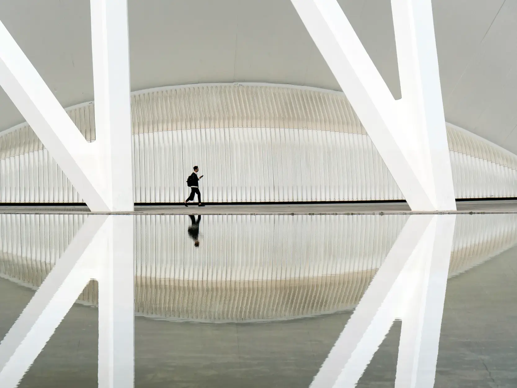 Ein Mann mit Rucksack geht durch die weiße Architektur der Ciutat de les Arts i les Ciències, die sich im Wasser spiegelt.