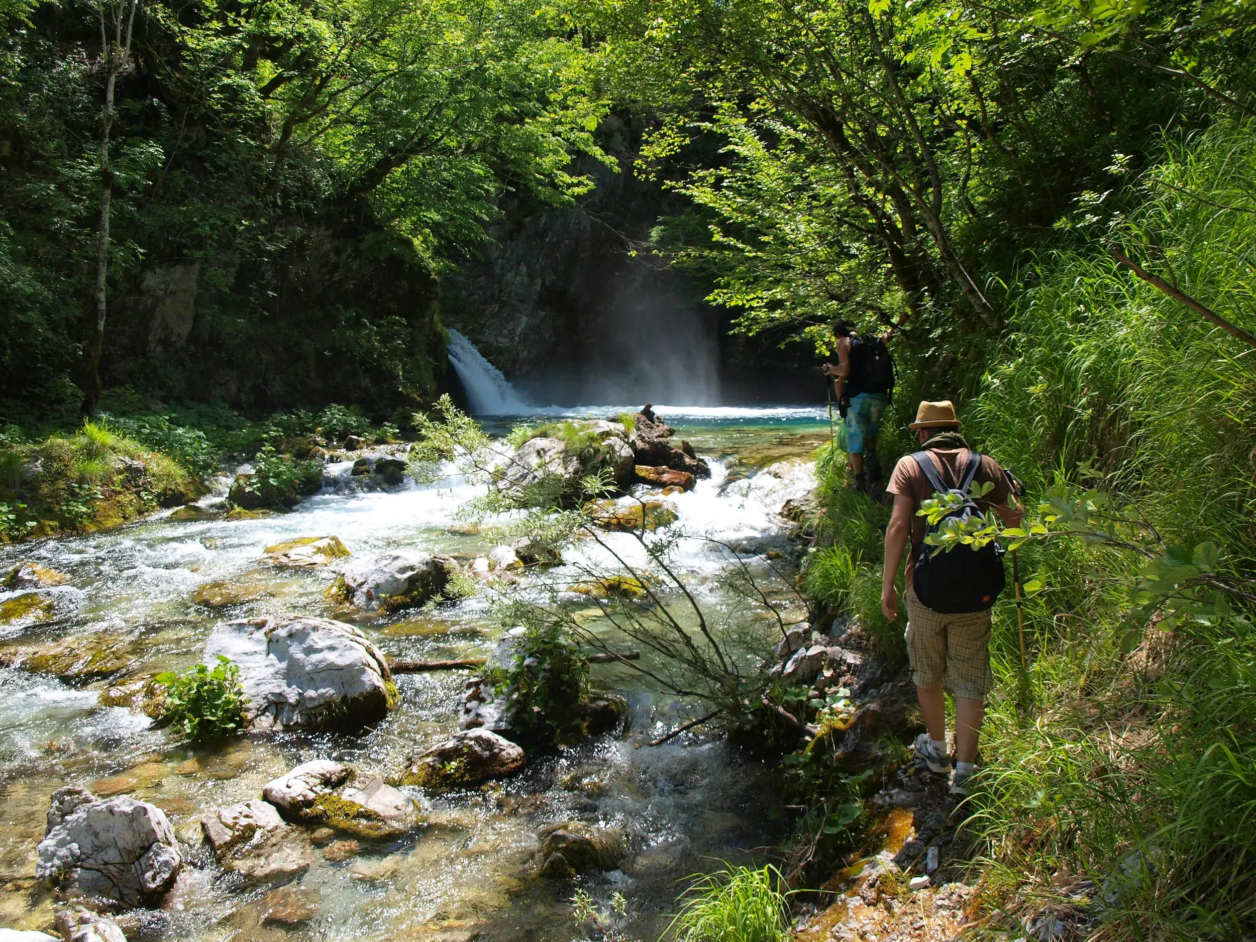 Zwei Wanderer auf einem schmalen Pfad am klaren Gebirgsfluss, der durch einen Wald zum Wasserfall 'Blaues Auge' in Albanien führt.