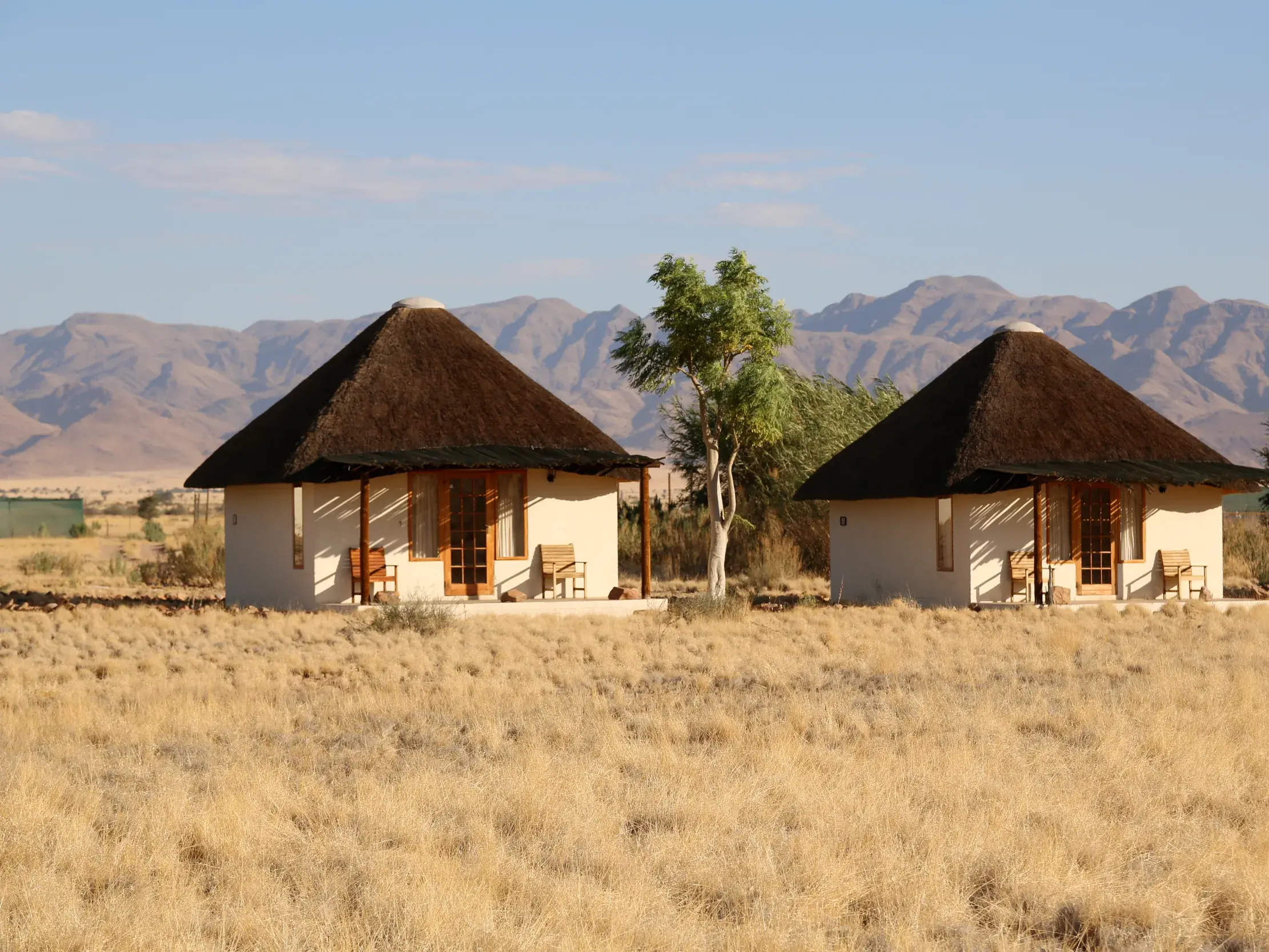 Zwei reetgedeckte Lodge-Bungalows in der weiten Savanne Namibias mit Blick auf die markanten Berge des Erongo-Gebirges.