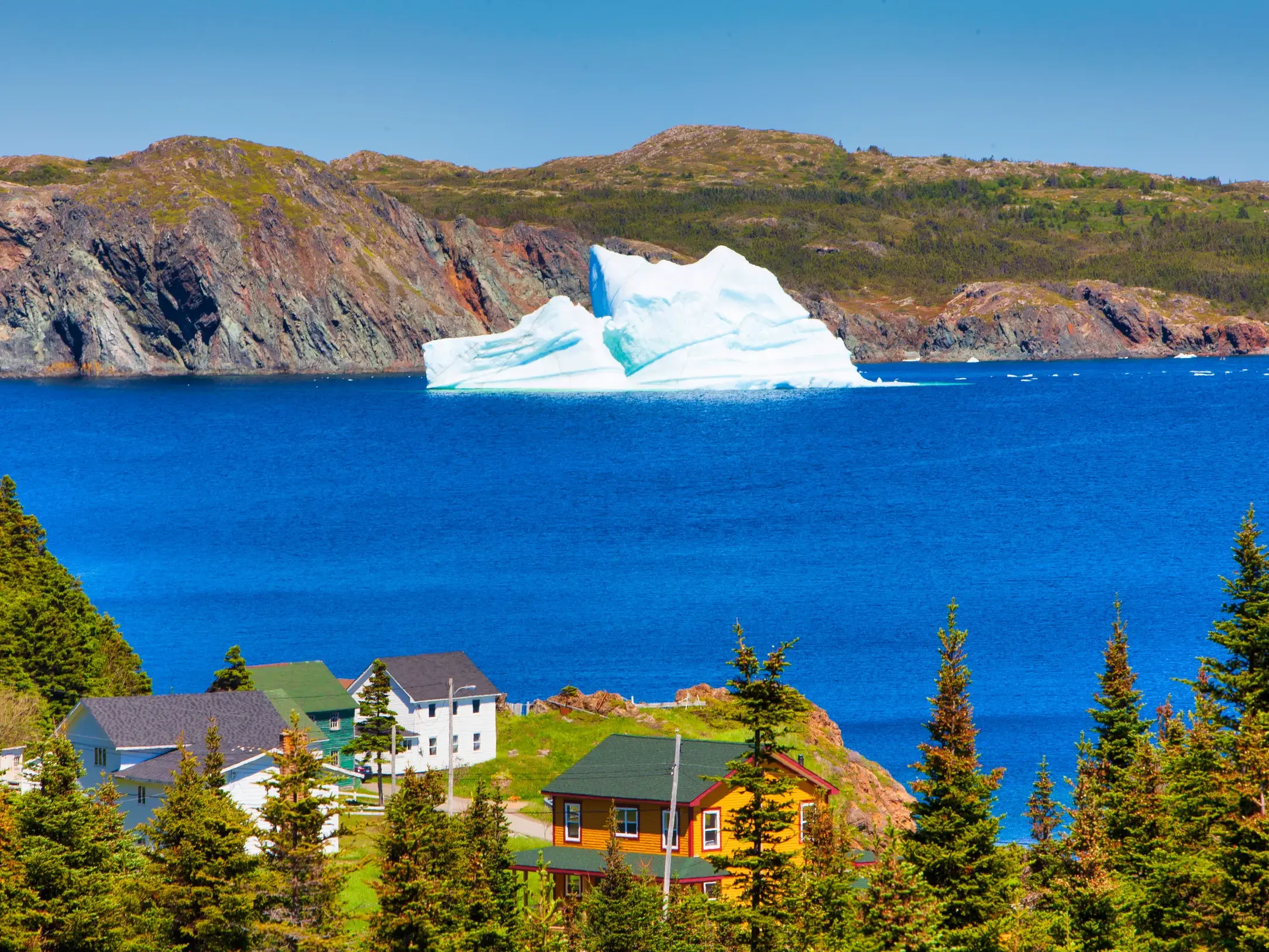 Majestätischer Eisberg treibt vor der Küste von Twillingate, Neufundland, mit bunten Häusern an der zerklüfteten Atlantikküste.