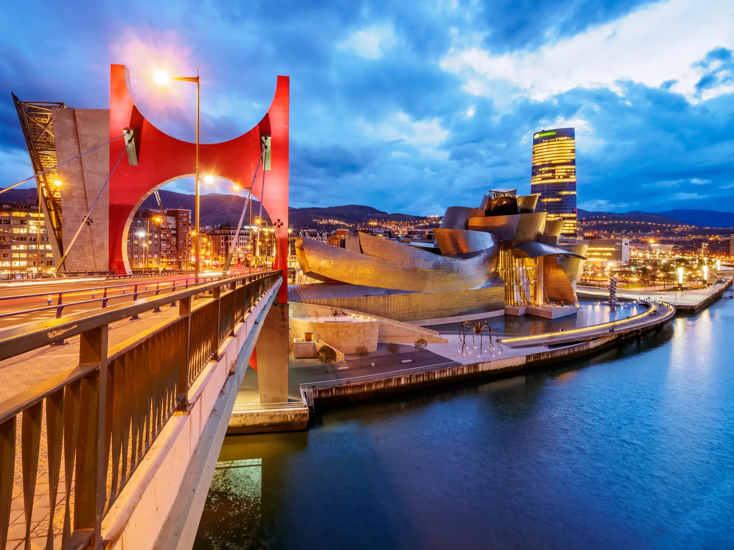 Das beleuchtete Guggenheim-Museum in Bilbao neben der roten La-Salve-Brücke am Fluss Nervión bei Dämmerung.