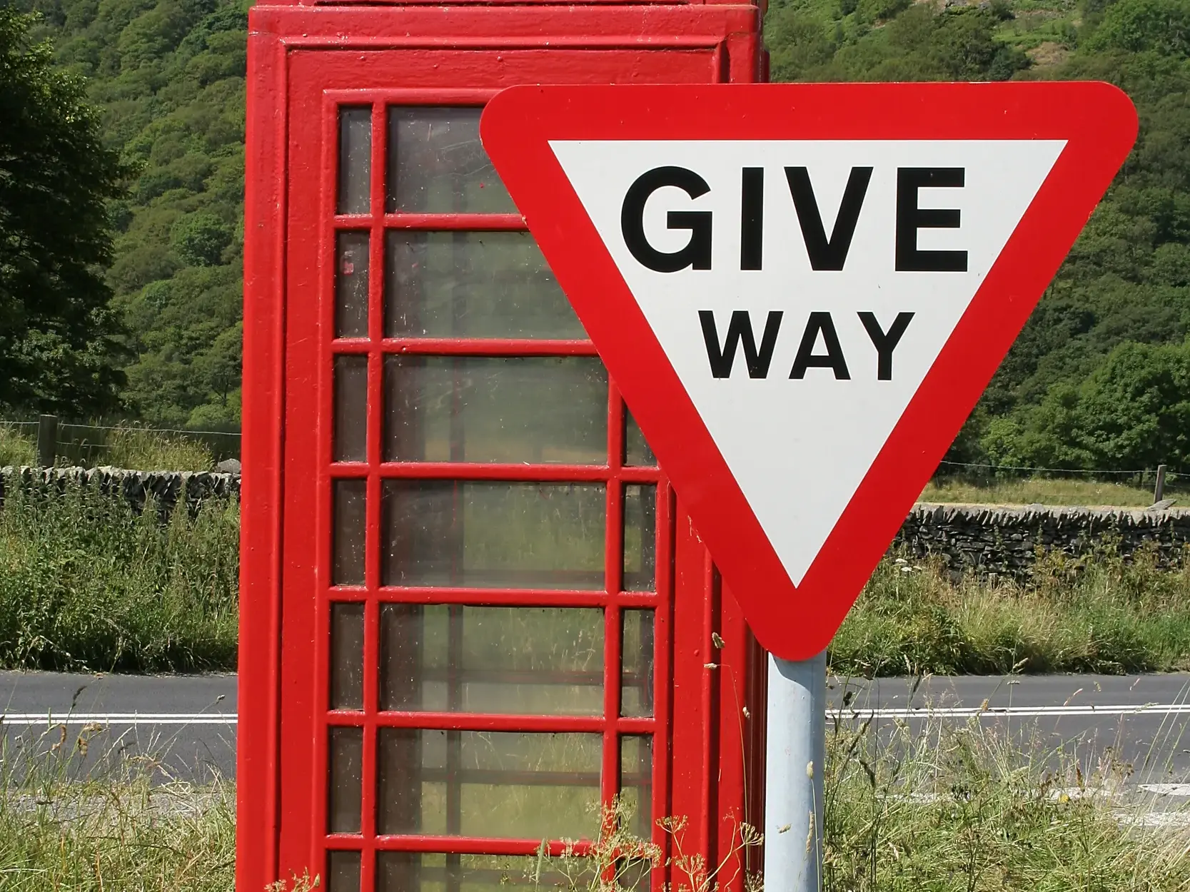 Eine klassische rote Telefonzelle und ein 'Give Way'-Verkehrsschild stehen an einer Landstraße vor der grünen Hügellandschaft der schottischen Highlands.