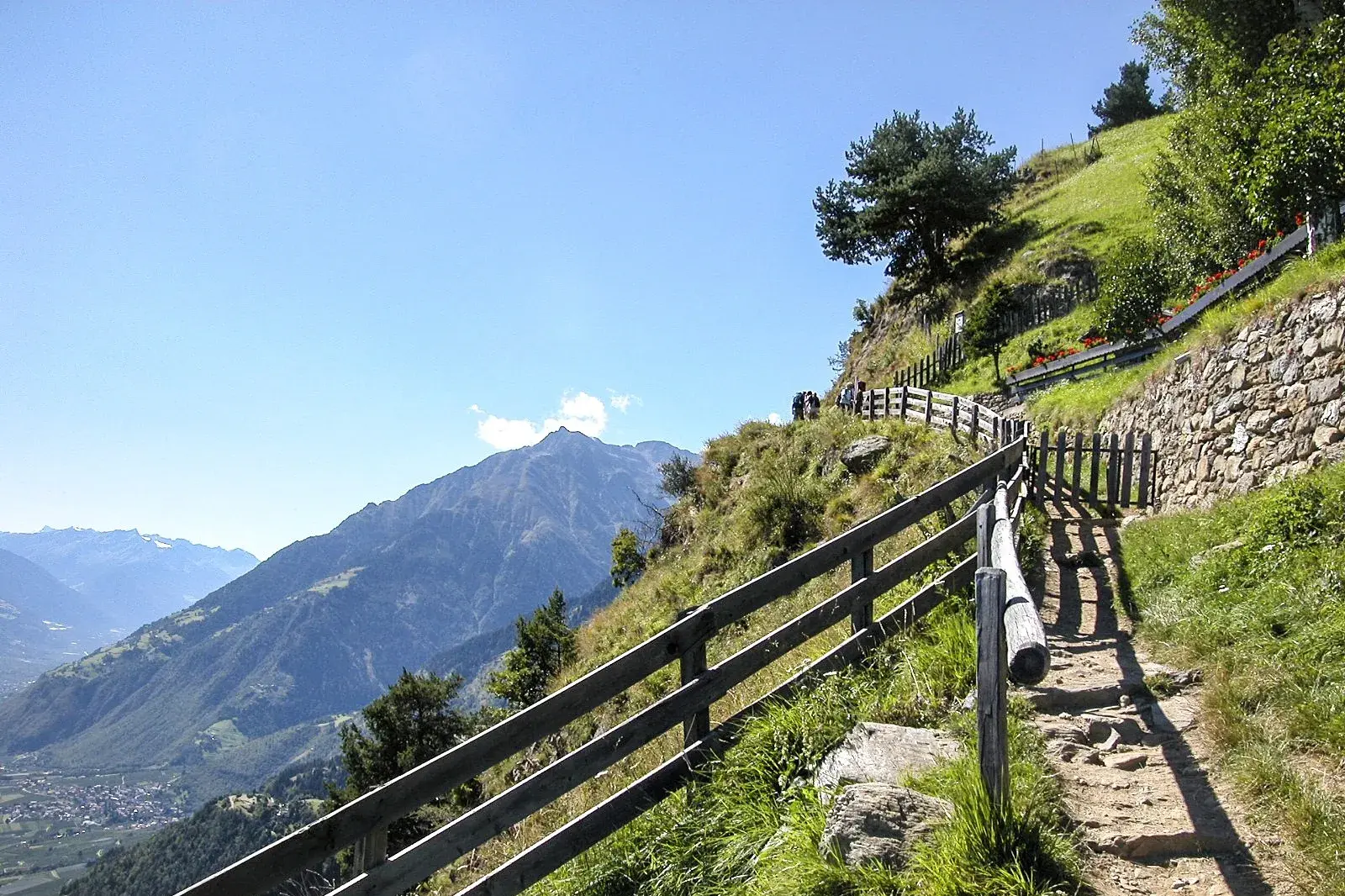 Ein sonniger Wanderweg mit Holzzaun führt hoch über dem Vinschgau entlang und bietet ein Panorama auf die Texelgruppe.