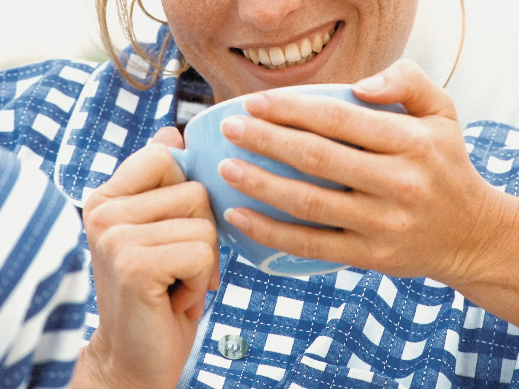 Eine lächelnde Frau in einem blau-weiß karierten Hemd genießt eine Fika und hält eine hellblaue Tasse fest.