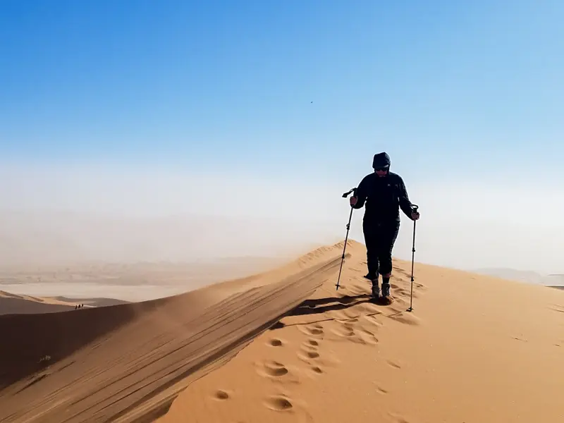 Wanderer mit Trekkingstöcken besteigt den windigen Kamm einer hohen Sanddüne im Dünenmeer von Sossusvlei, Namibia.