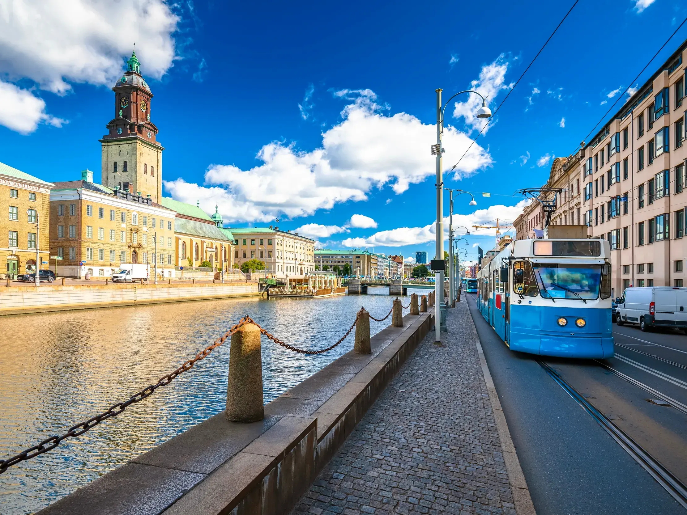 Eine ikonische blaue Straßenbahn fährt am Stora Hamnkanalen in Göteborg vorbei, im Hintergrund der Turm der Deutschen Kirche.