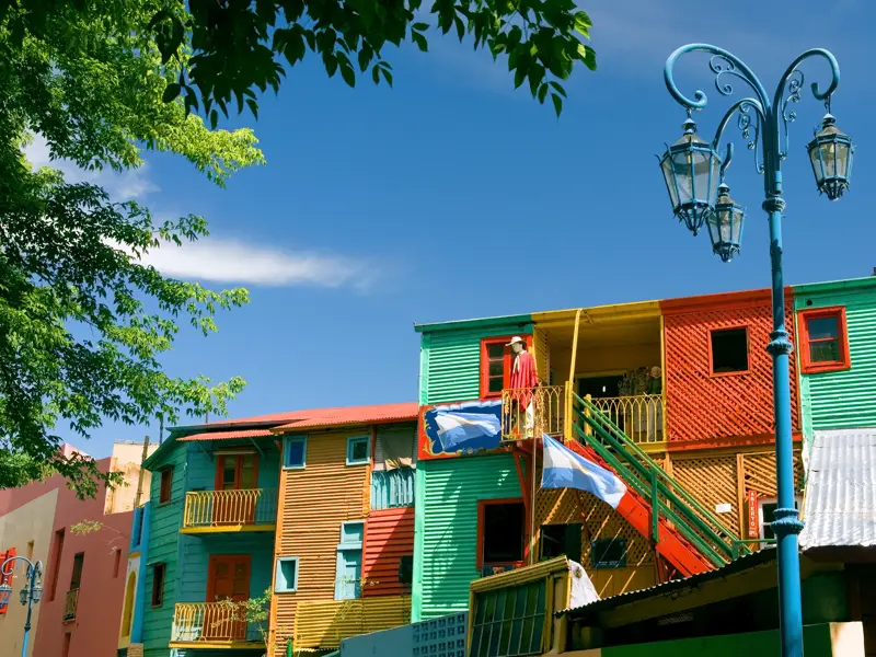 Knallbunte Wellblechhäuser in La Boca, Buenos Aires, mit argentinischer Flagge an einem Balkon bei strahlend blauem Himmel.