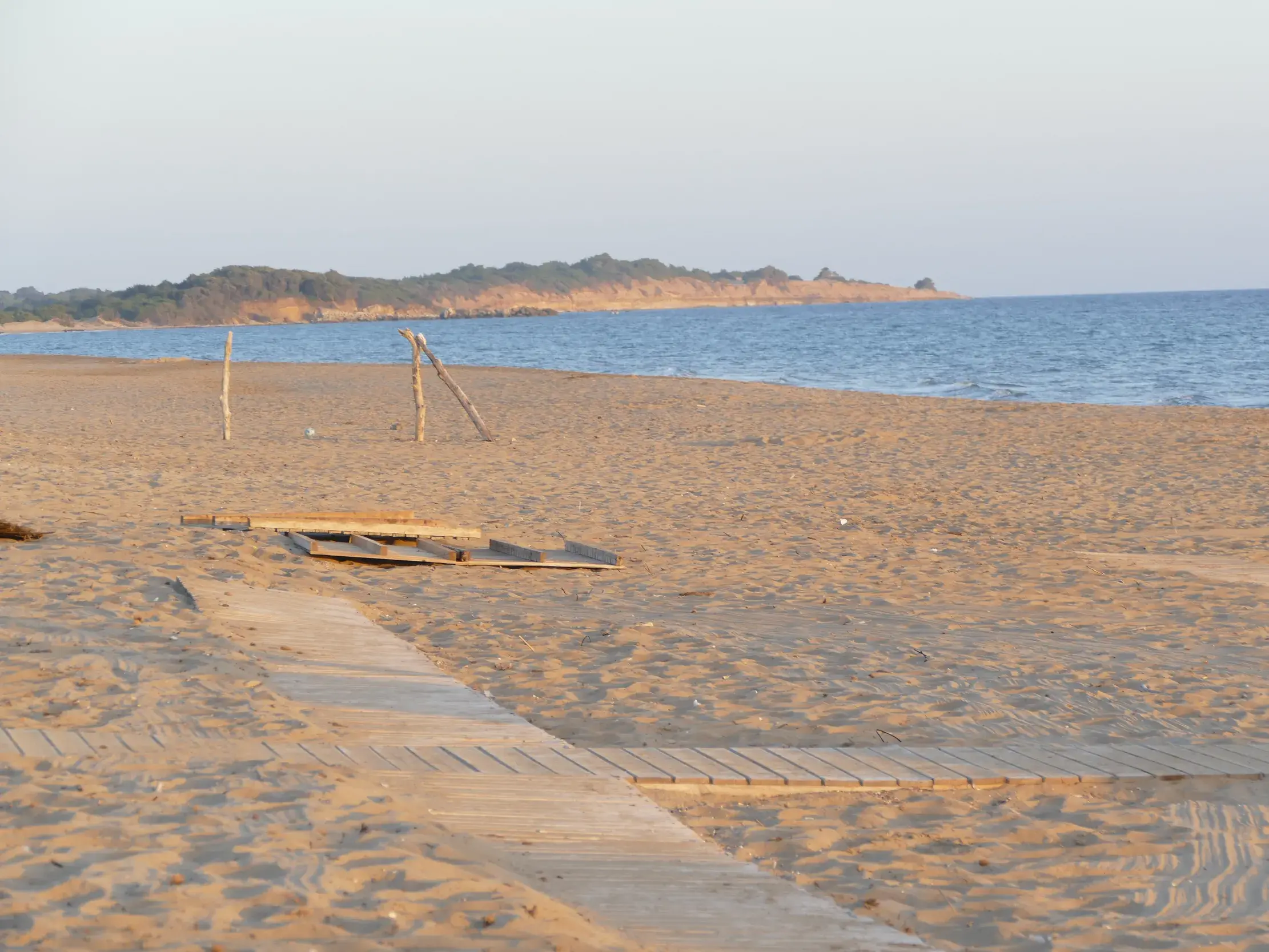 Ein Holzpfad führt über den weiten Sandstrand von Marathias auf Korfu, im Hintergrund das Meer und die Küstenlinie.