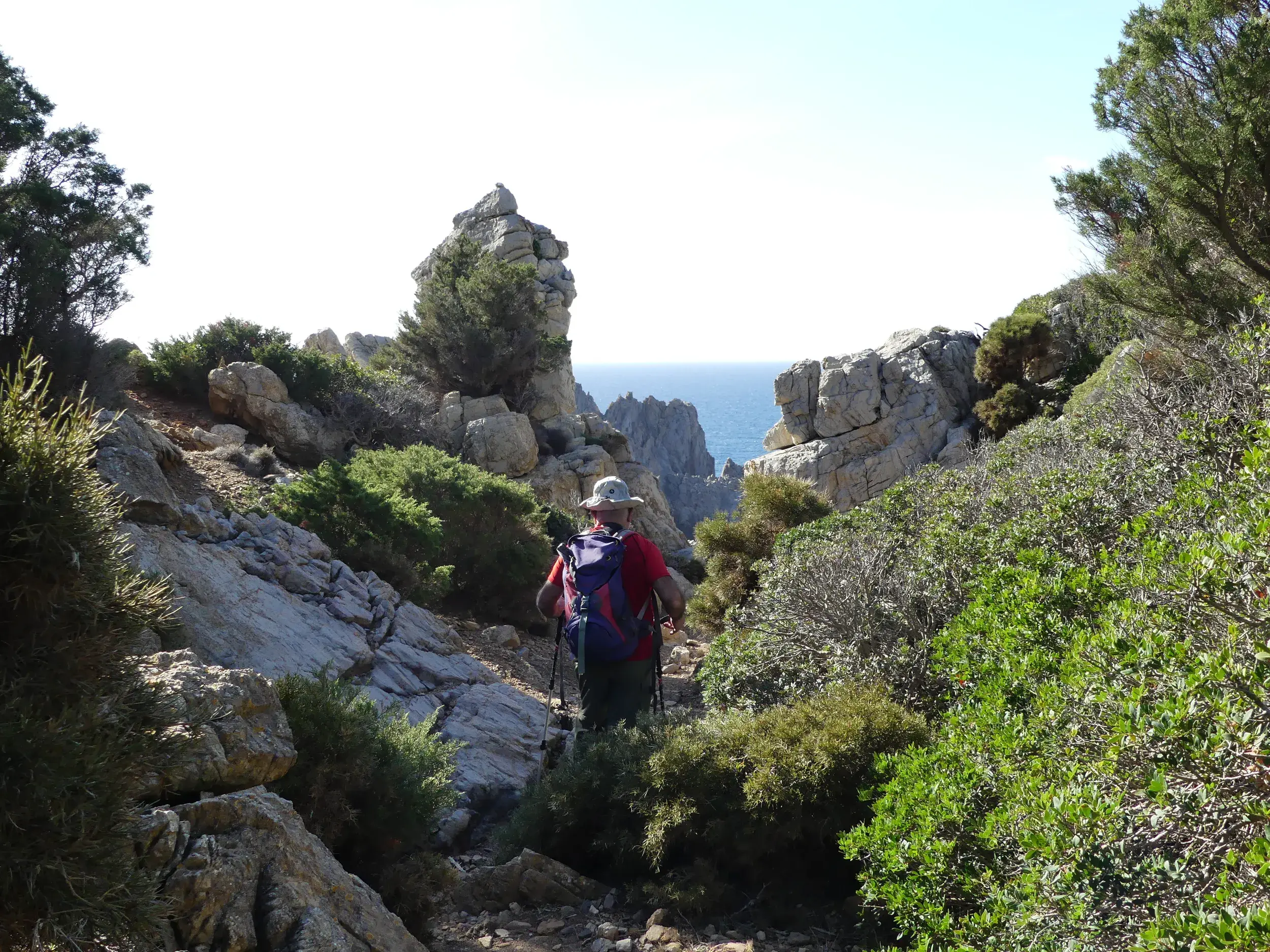 Wanderer mit Rucksack auf einem Küstenpfad am Capo Pecora auf Sardinien, umgeben von bizarren Granitfelsen und mit Blick aufs Meer.