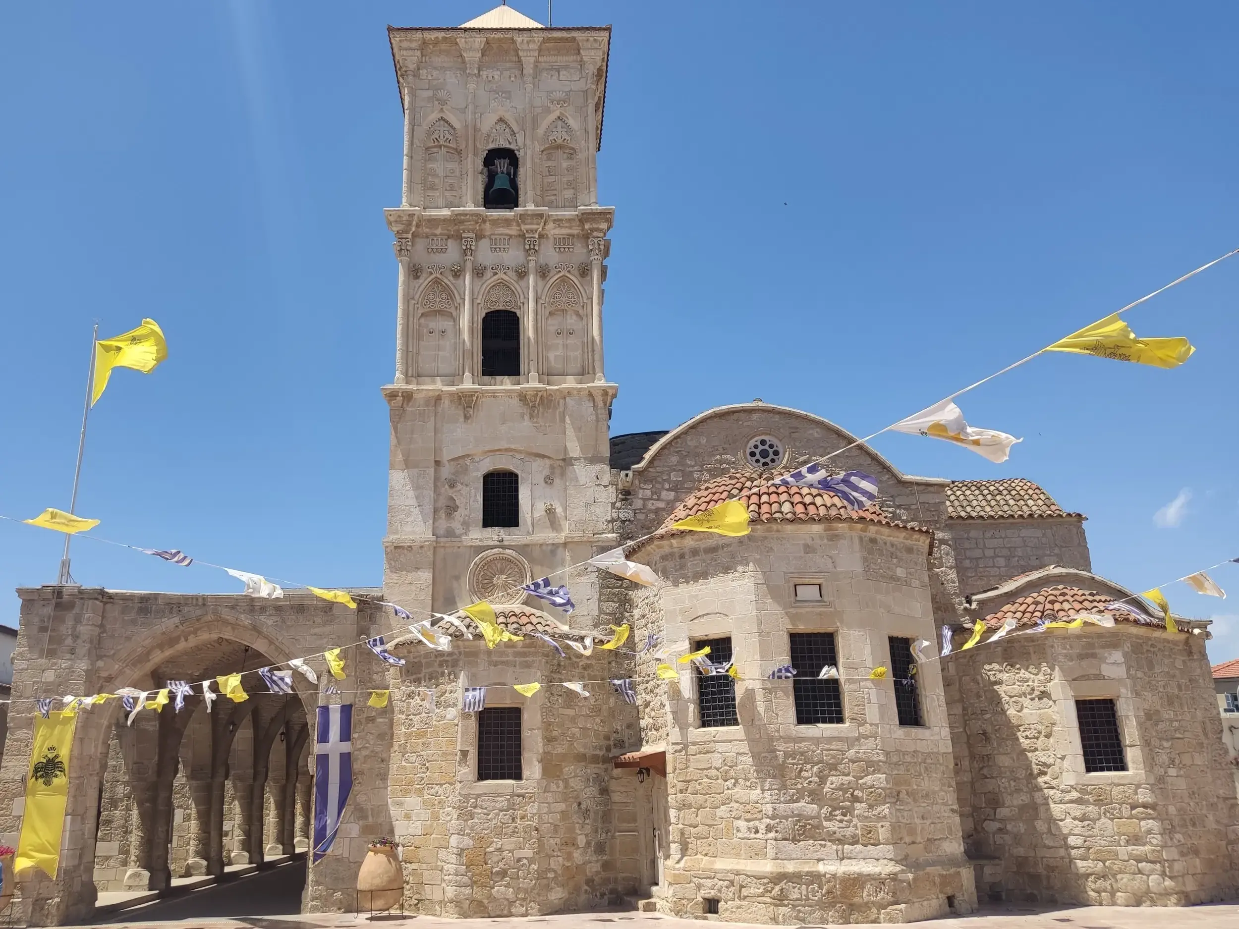 Die imposante Lazaruskirche in Larnaka aus hellem Stein, mit Arkadengang und wehenden Flaggen an einem sonnigen Tag auf Zypern.