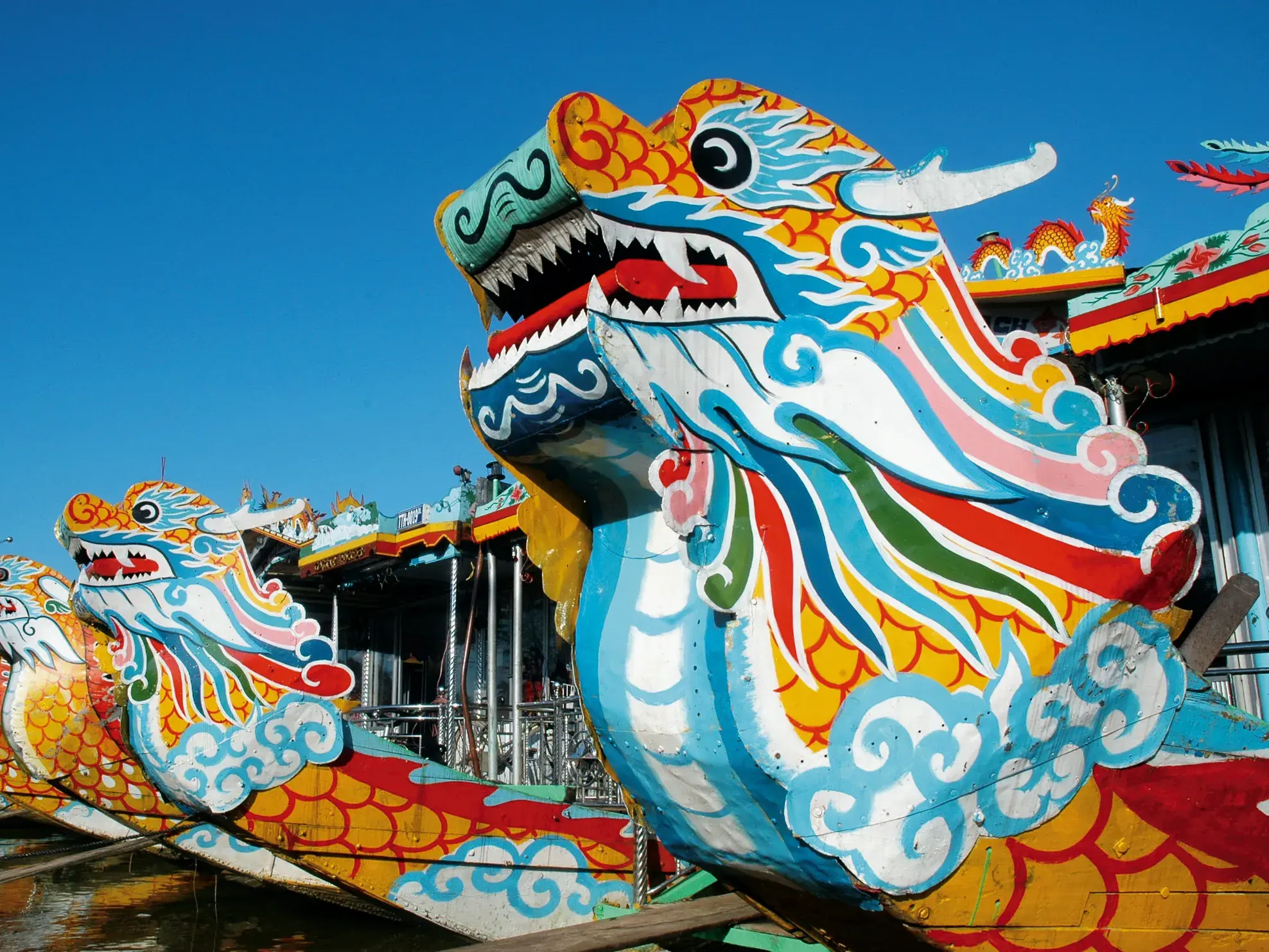 Farbenfrohe, traditionelle Drachenboote liegen aufgereiht am Ufer des Parfümflusses in Hue unter blauem Himmel.