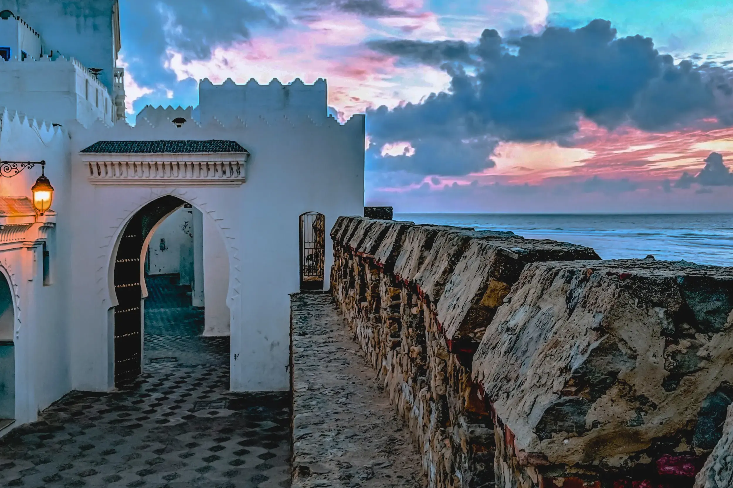 Weg entlang der steinernen Stadtmauer von Asilah mit Blick auf den Atlantik unter einem farbenfrohen Abendhimmel.