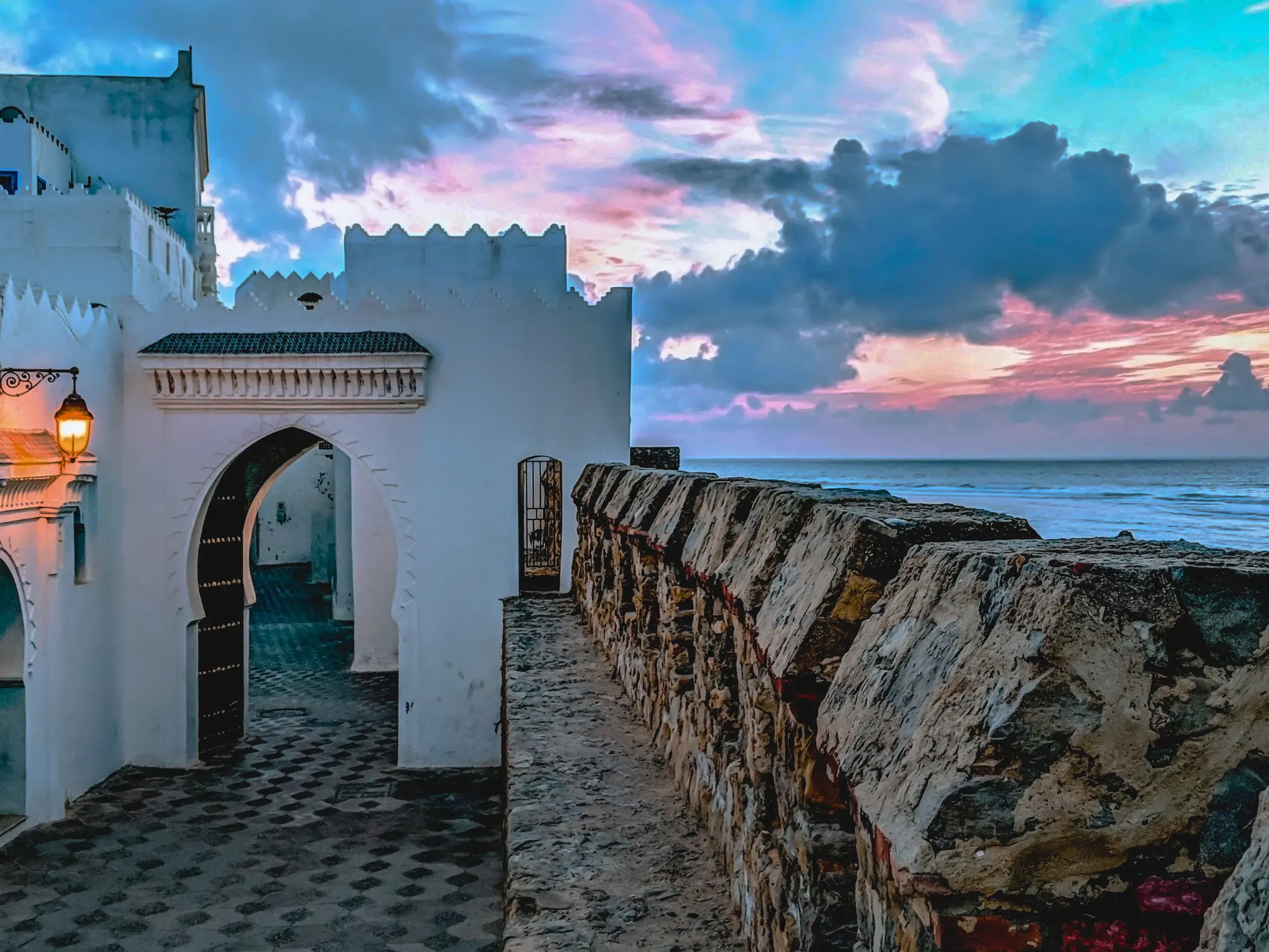Weg entlang der steinernen Stadtmauer von Asilah mit Blick auf den Atlantik unter einem farbenfrohen Abendhimmel.