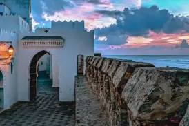 Weg entlang der steinernen Stadtmauer von Asilah mit Blick auf den Atlantik unter einem farbenfrohen Abendhimmel.