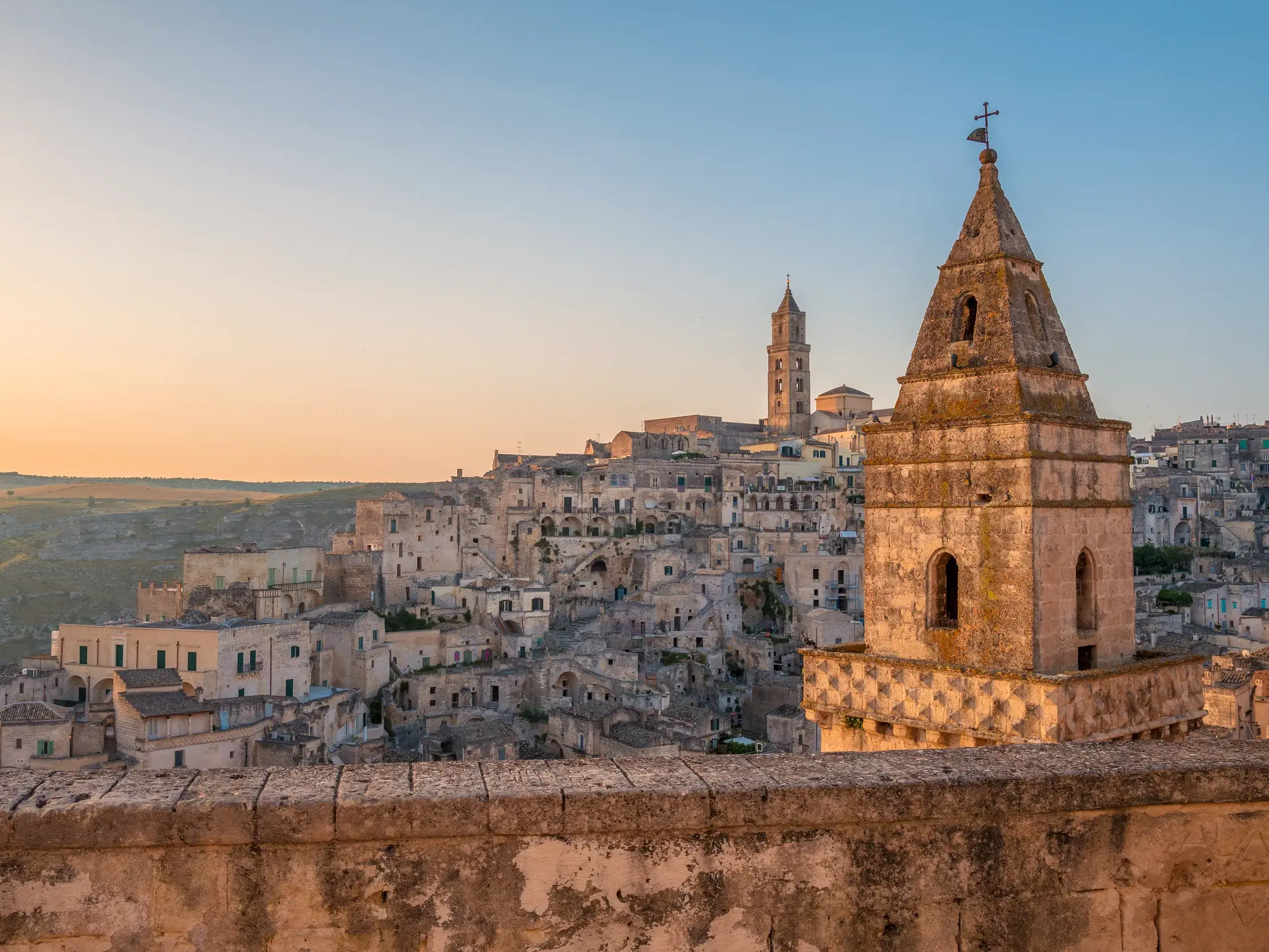 Ein alter Kirchturm im Vordergrund mit Blick auf die historischen Höhlenwohnungen der Sassi in Matera im warmen Morgenlicht.