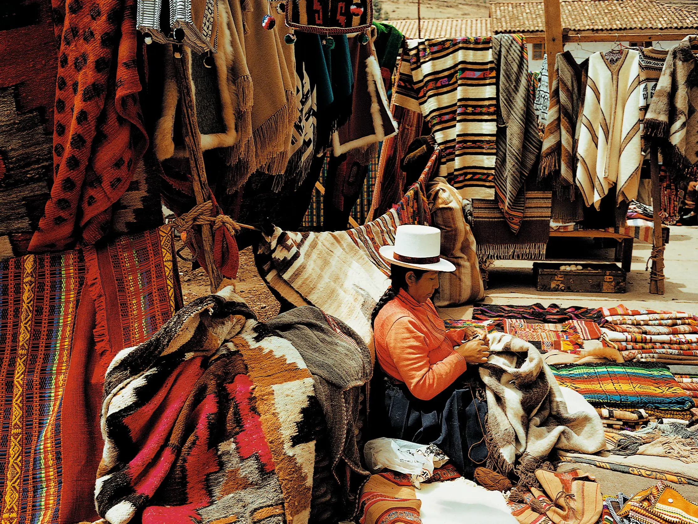 Eine Weberin in Tracht fertigt auf dem Markt von Chinchero in Peru handgemachte Textilien zwischen bunten Decken und Ponchos.