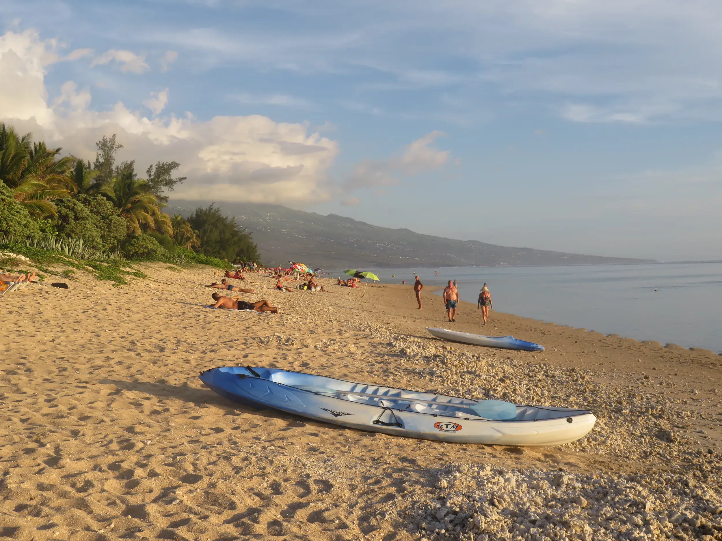 Ein blau-weißes Kajak liegt am sonnigen Sandstrand von La Saline les Bains, während Menschen am Meer auf La Réunion entspannen.